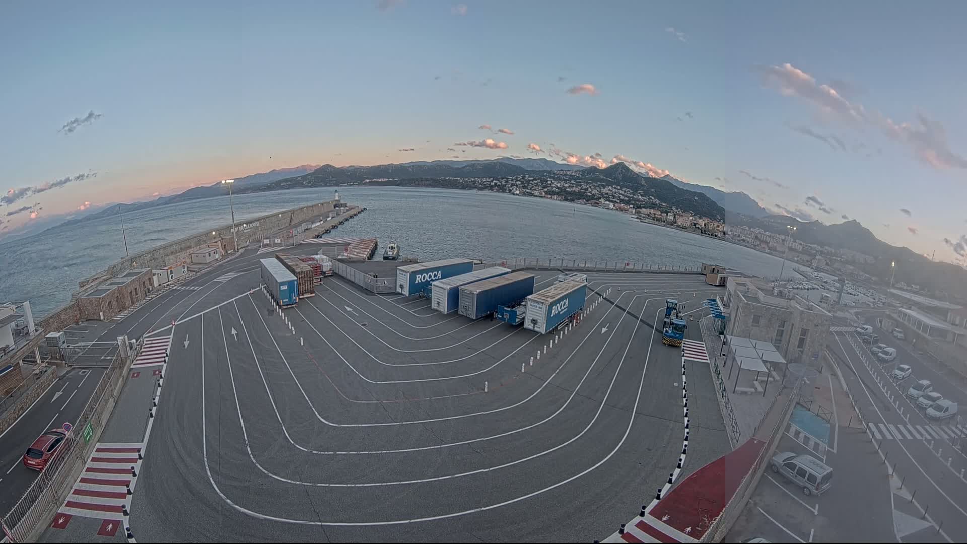 A port area with several semi-trailer trucks parked in a designated area, overlooking calm water and mountains under a clear, twilight sky.