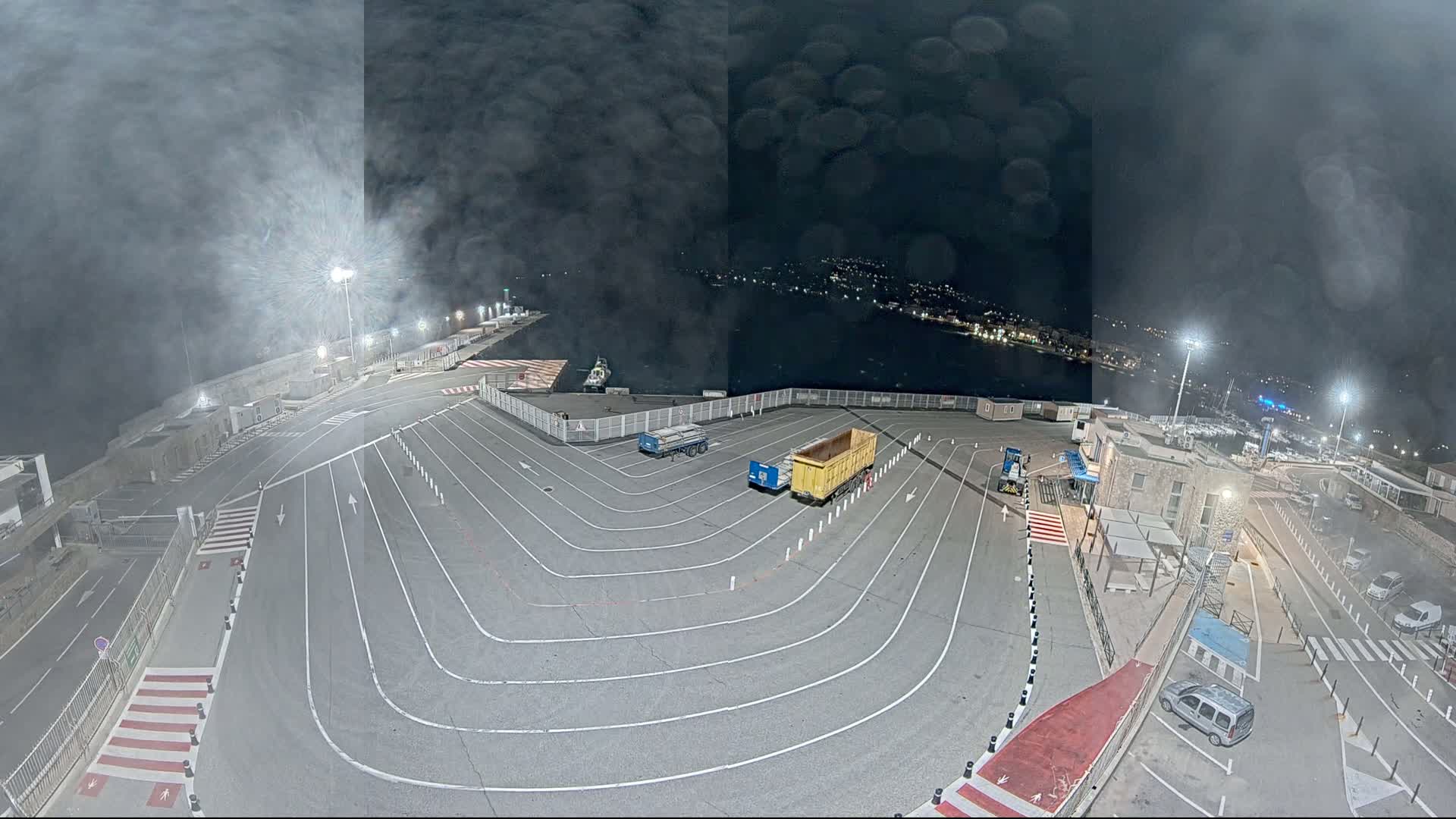 A nighttime, hazy view of a harbor's empty parking lot with two trucks and a few cars, near a waterfront with lights and buildings.