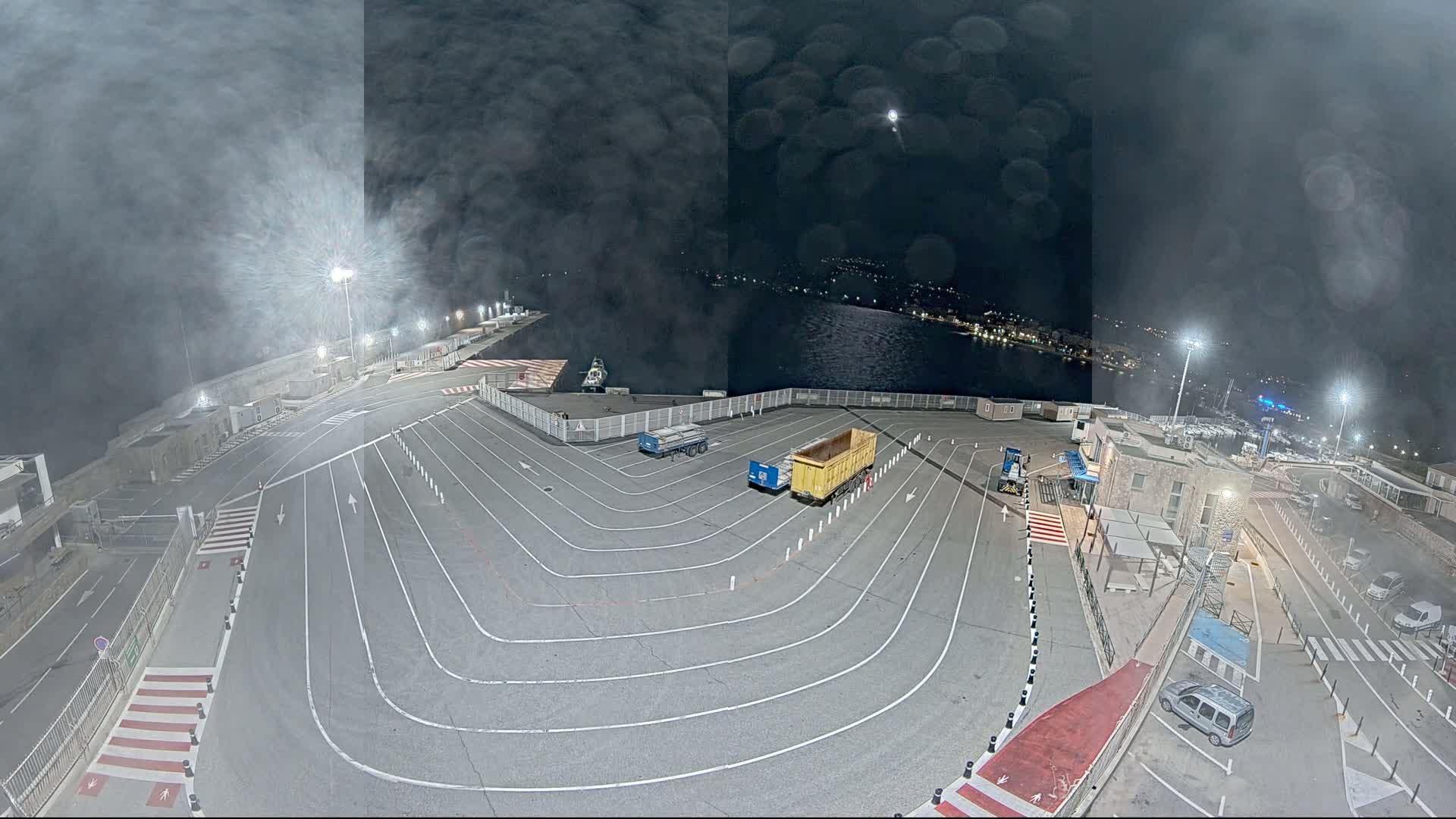 A nighttime aerial view of an empty port area with two trucks and several cars parked, under a hazy, cloudy sky.
