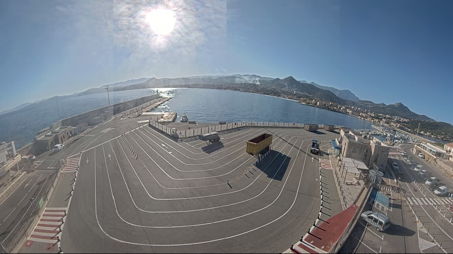 A sunny day overlooking a harbor with a large, empty paved area marked with driving lanes, several vehicles, and a town in the background surrounded by mountains.
