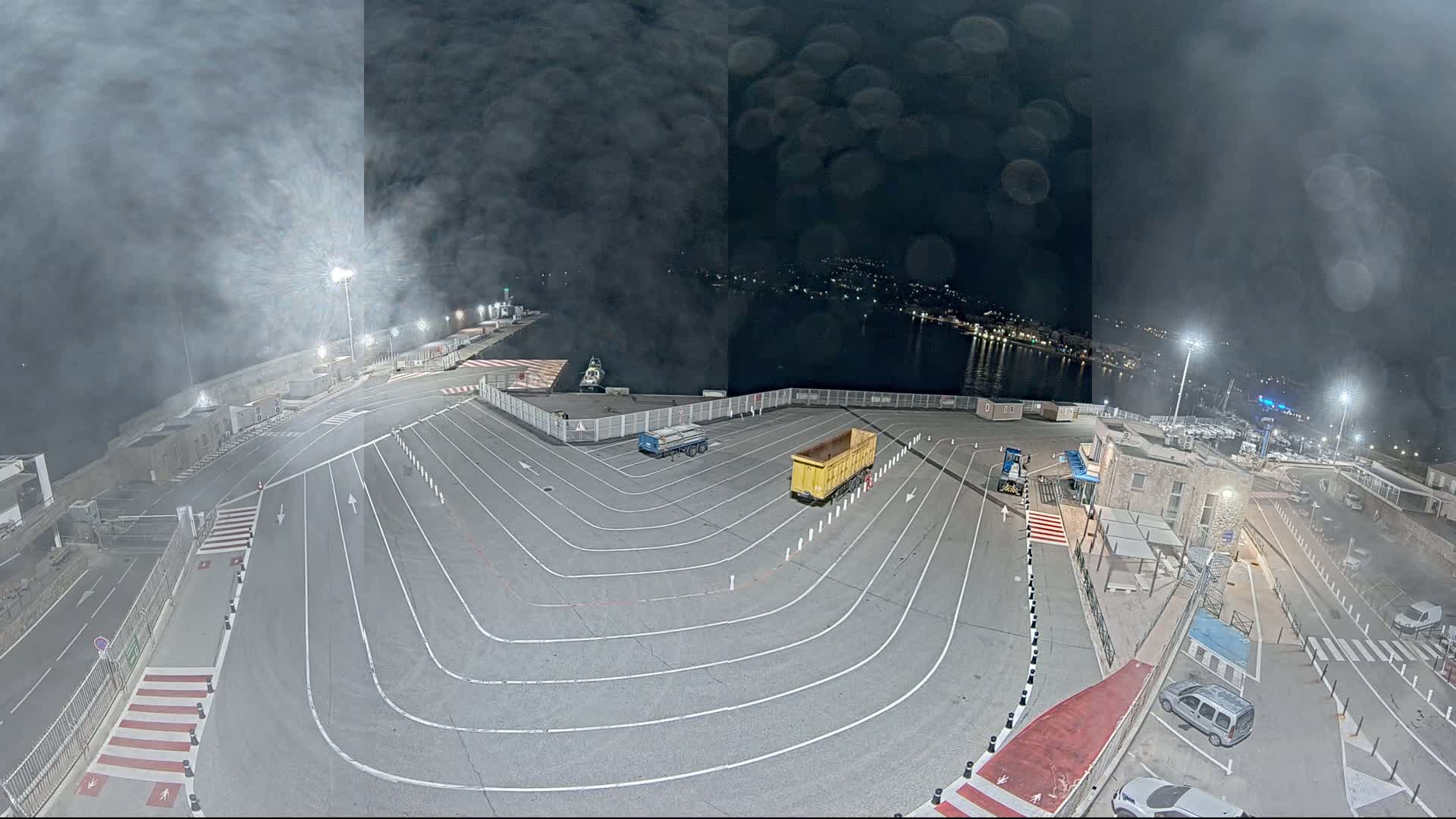 A nighttime, hazy view of a port's empty vehicle staging area with two trucks and a few cars, and a body of water with lights in the distance.