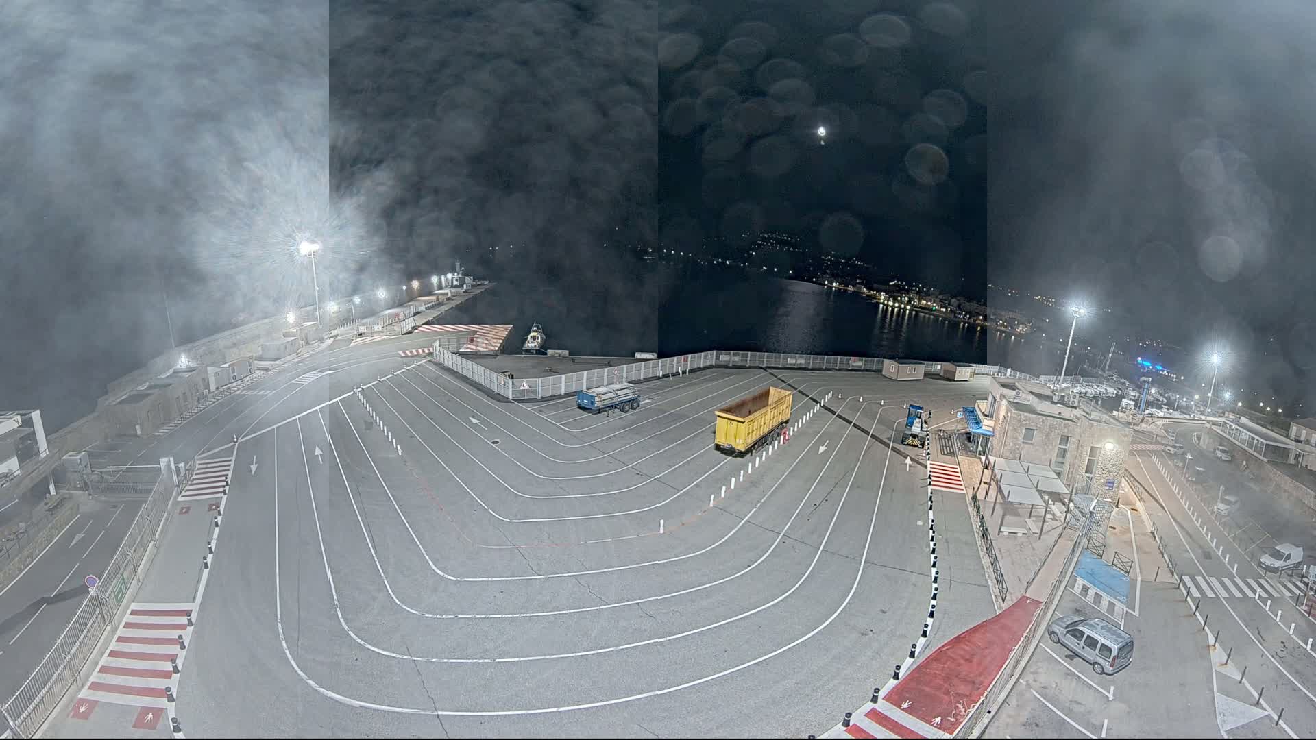 A nighttime aerial view of a harbor's loading area with two trucks parked on a marked asphalt surface, under a hazy, moonlit sky.