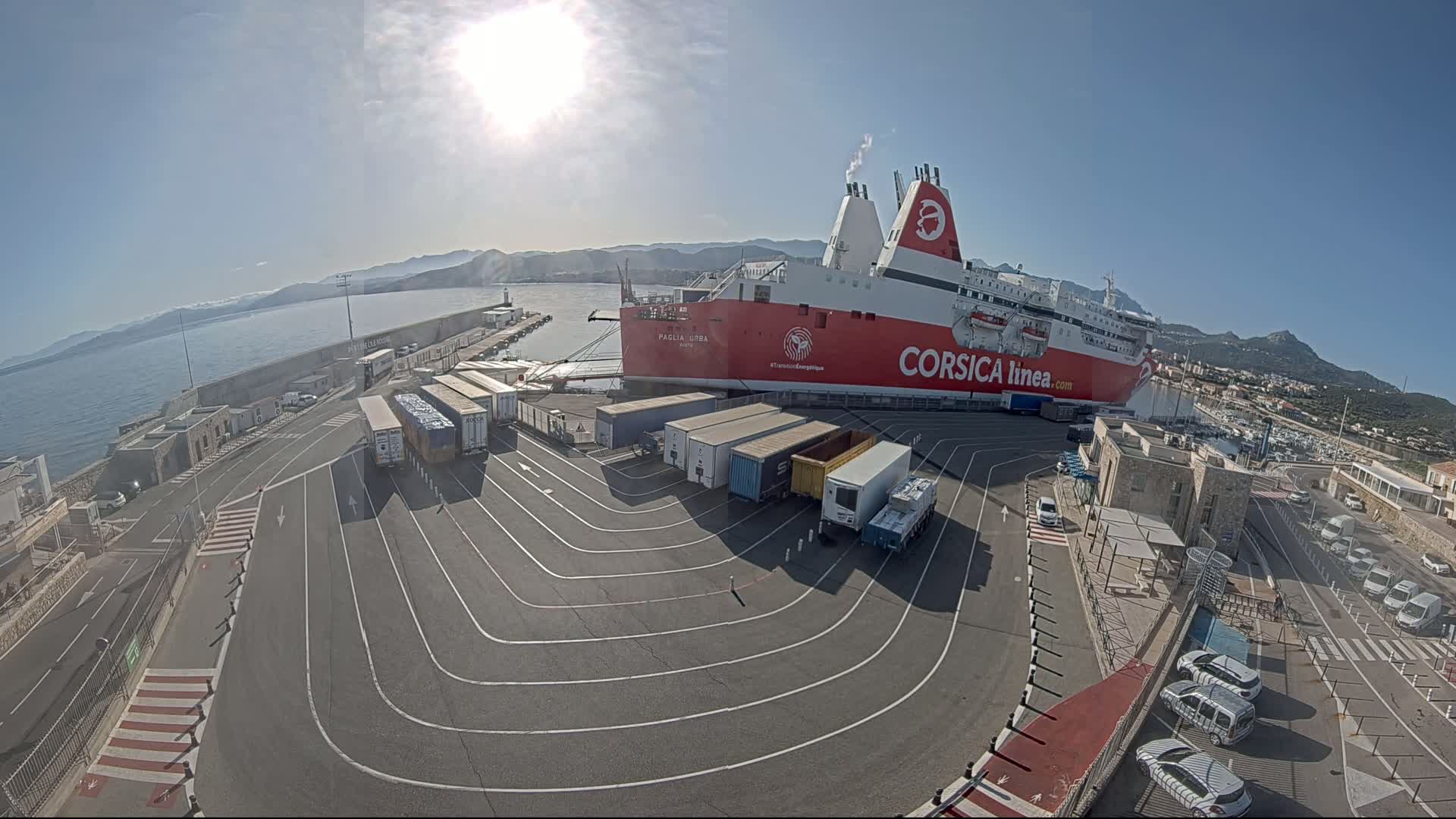 A large red and white ferry is docked at a port on a sunny day, with numerous trucks and cars parked nearby on a paved area next to calm blue water and mountains in the distance.
