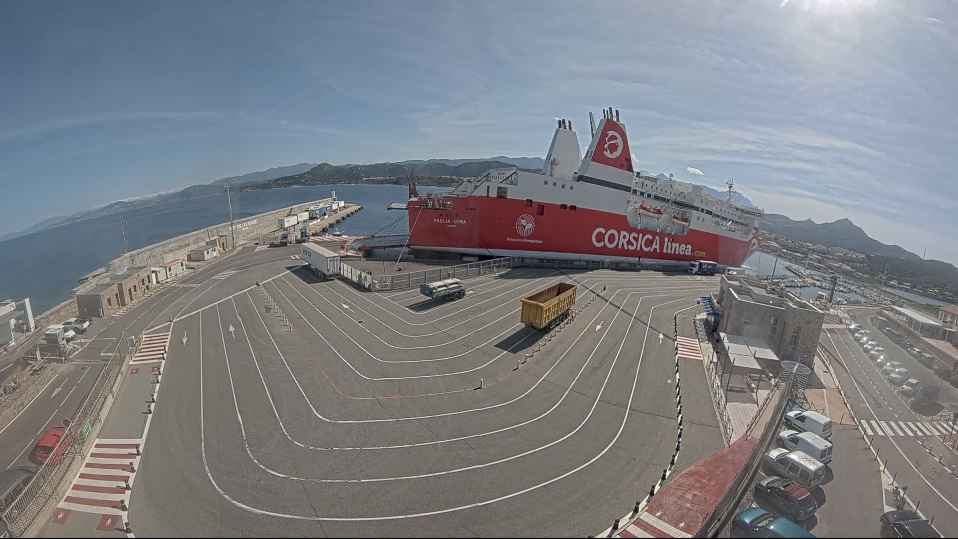 A large red and white ferry is docked at a port on a sunny day, with numerous vehicles parked nearby and curved lane markings guiding traffic around the vessel.