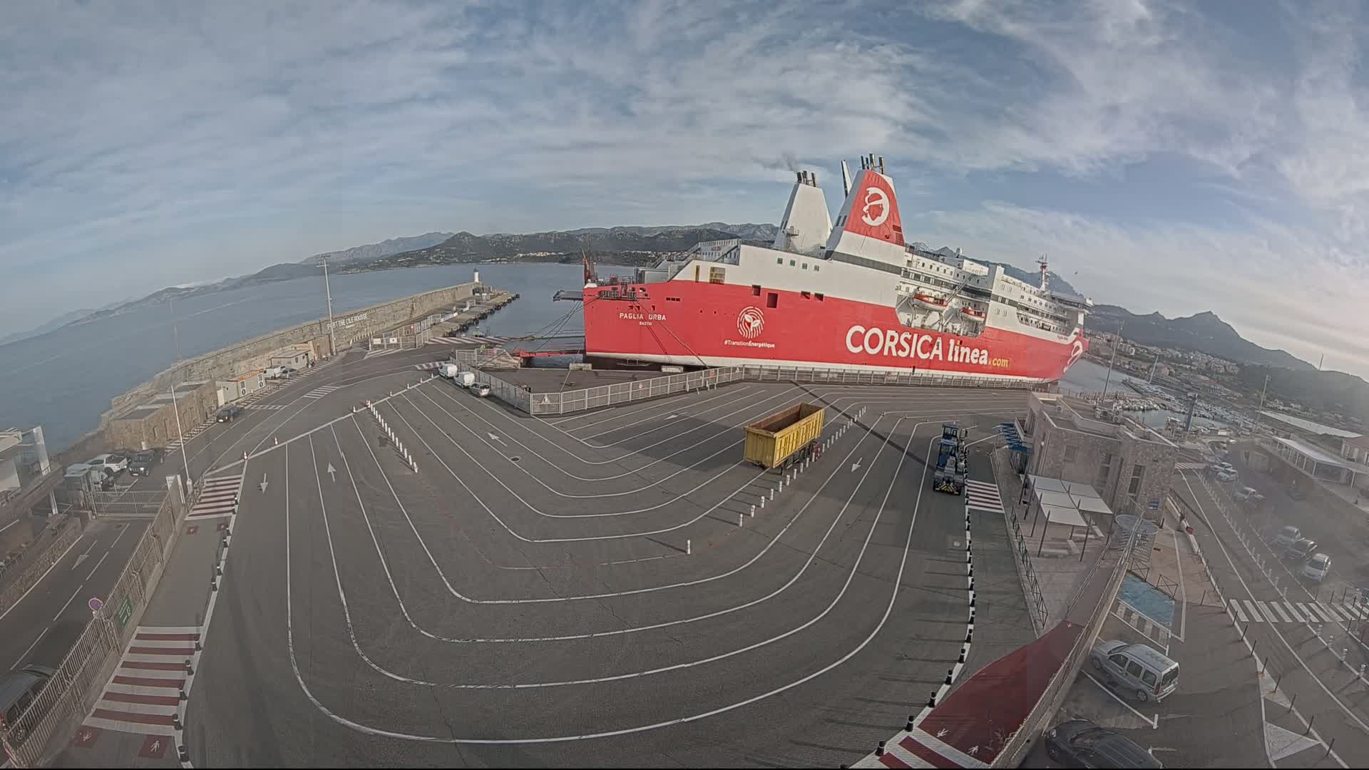 A large red and white ferry is docked at a port under a partly cloudy sky, with a paved area marked with driving lanes leading to it.