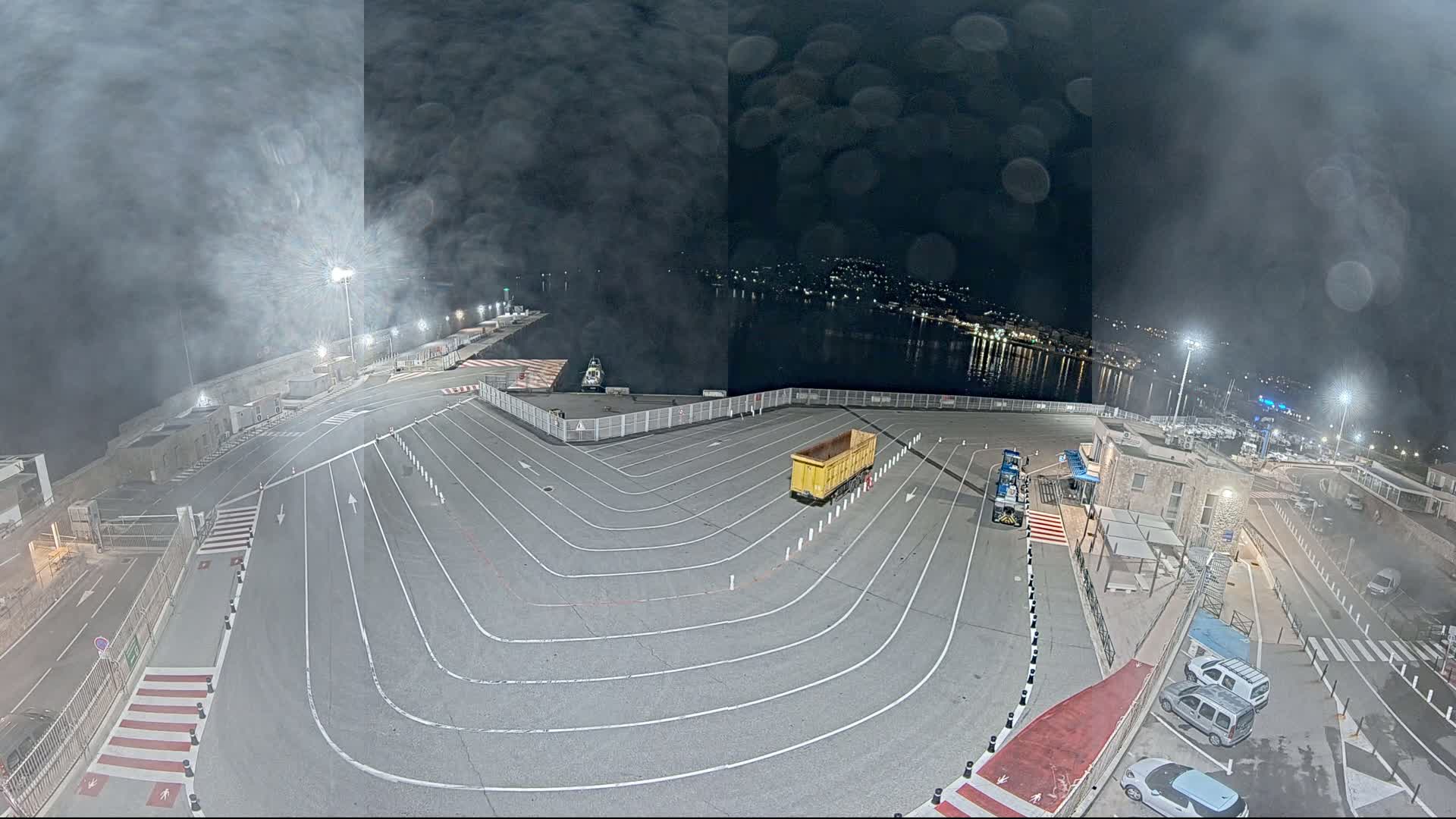 A nighttime aerial view of a port's empty parking lot with a yellow trailer, a few cars, and a hazy, possibly foggy, atmosphere.
