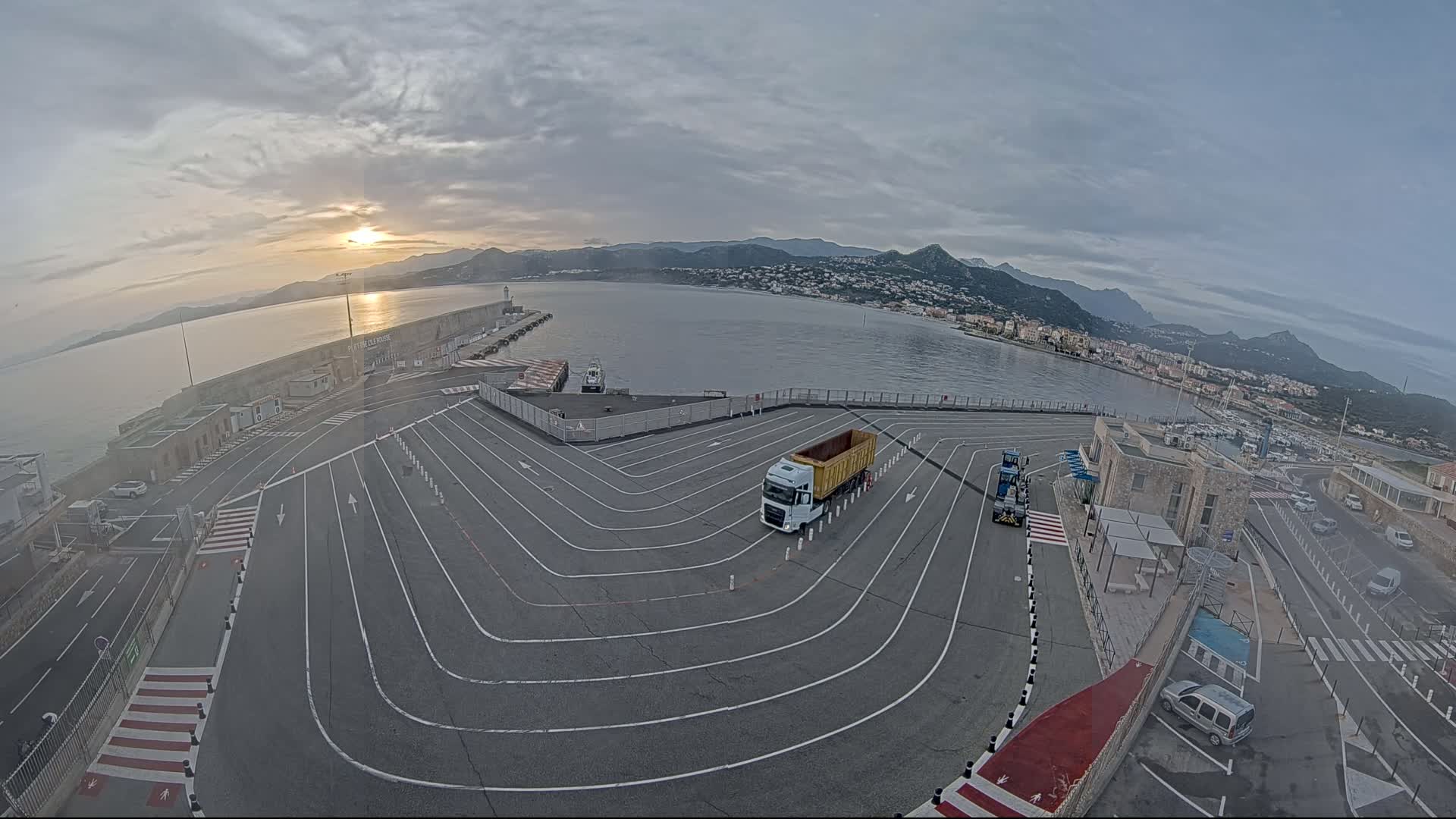 A semi-truck sits in a paved lot at a harbor, with a calm body of water and mountains in the background under a partly cloudy sunrise.