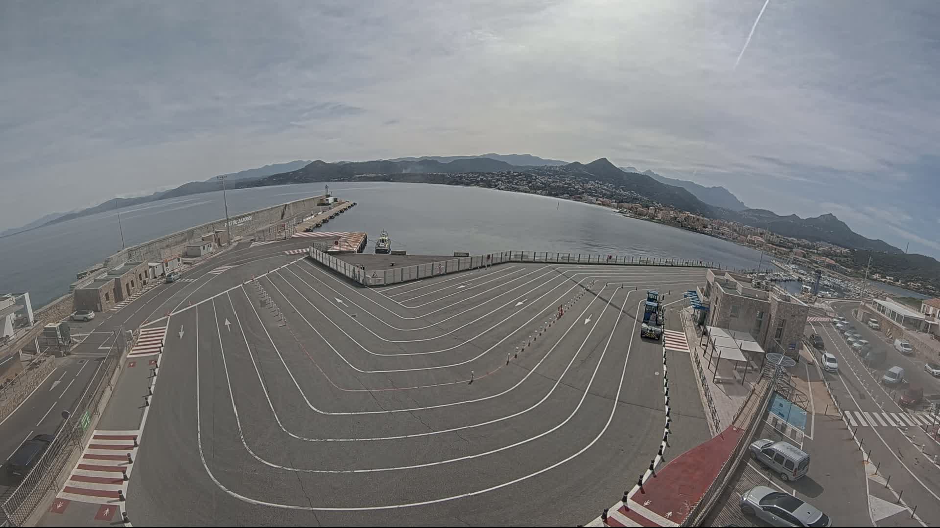 A ferry terminal with marked lanes for vehicles overlooks a calm bay and distant mountains under a mostly cloudy sky.