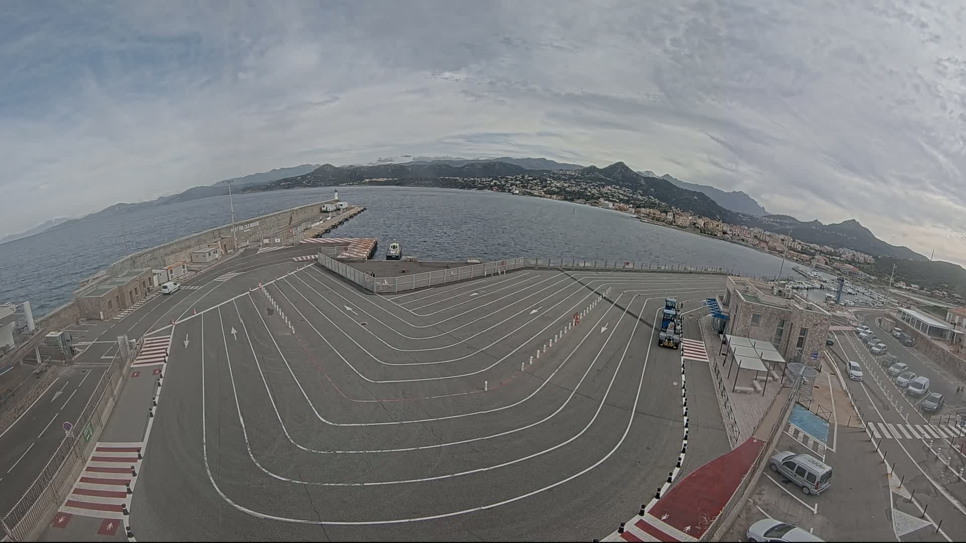An aerial view of a ferry terminal with marked parking lanes, overlooking a calm body of water and distant mountains under an overcast sky.