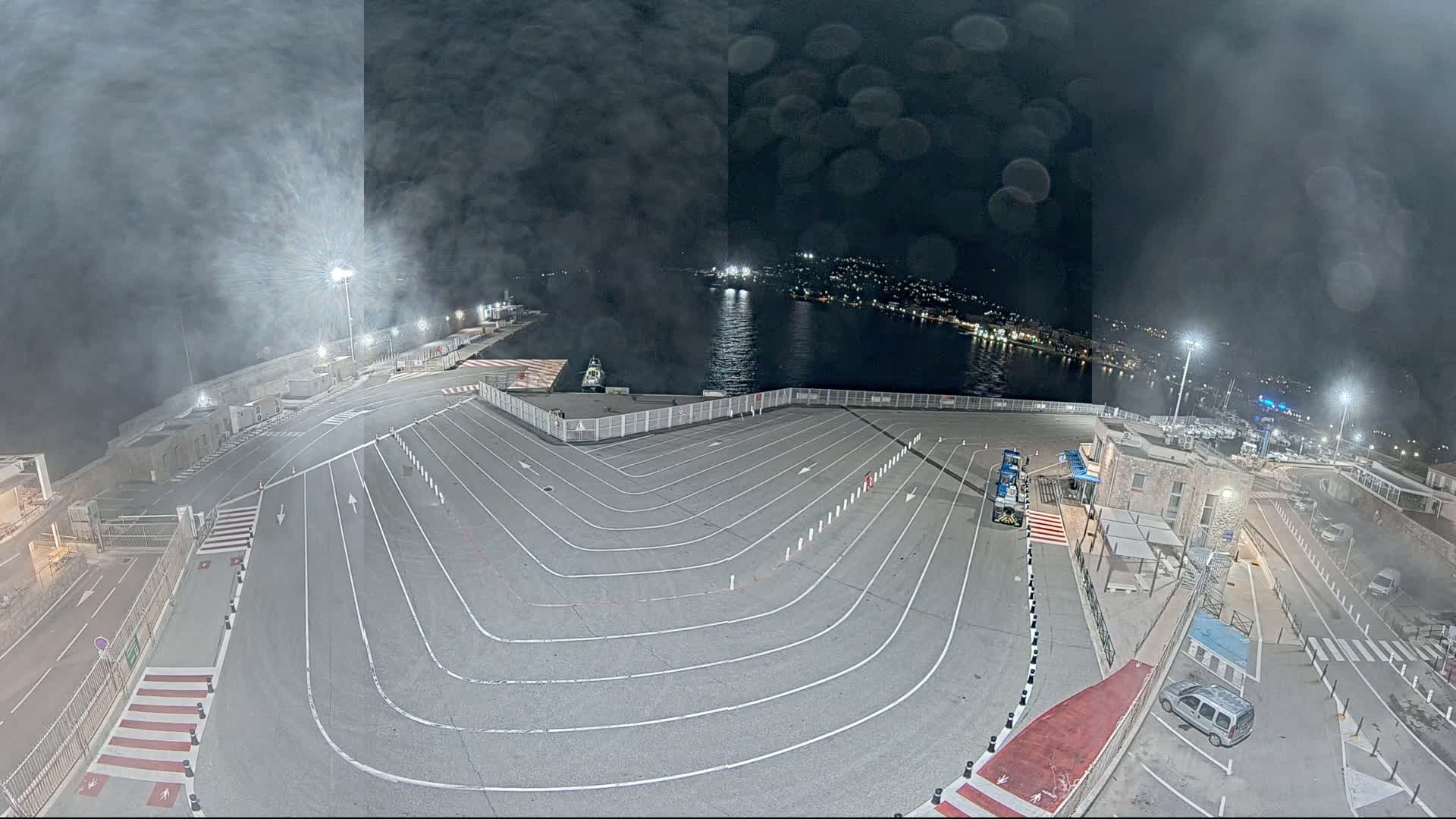 A nighttime aerial view of a ferry terminal with marked lanes and a body of water visible in the background under hazy conditions.