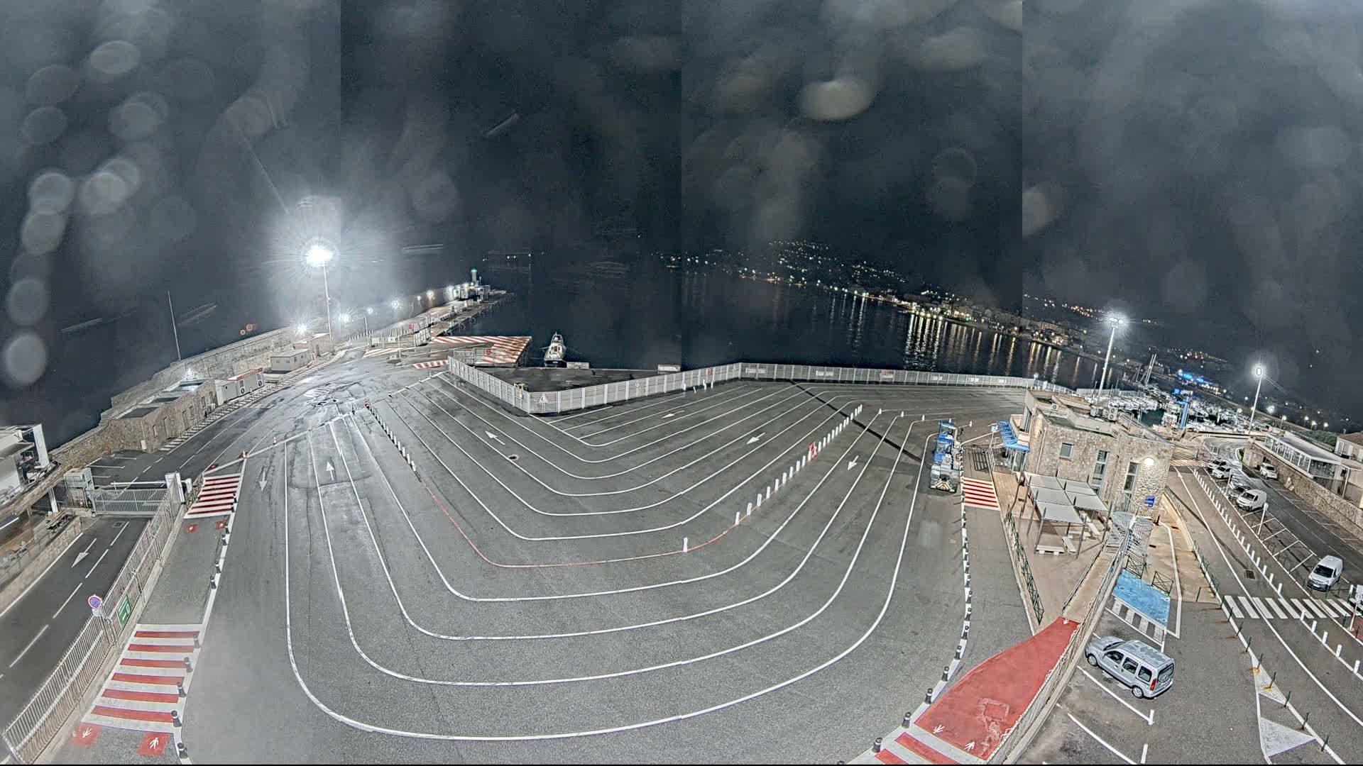 A nighttime, wet view of an empty ferry terminal with marked lanes and a few cars parked nearby, overlooking a calm body of water with lights from a town visible in the distance.