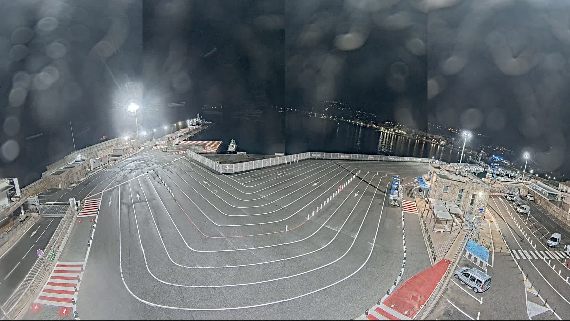 A nighttime, rain-streaked view of a ferry terminal with marked vehicle lanes, a small harbor, and parked cars.