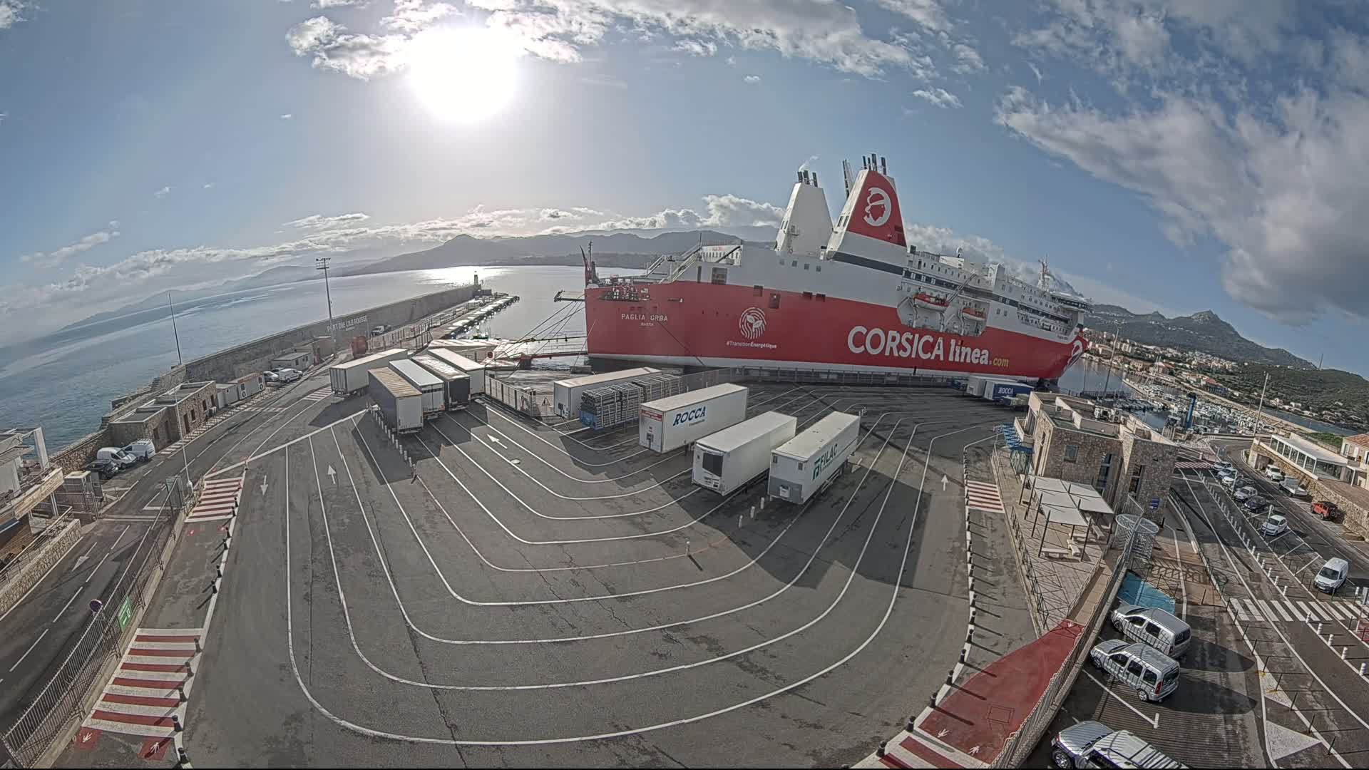 A large red and white ferry is docked at a port on a sunny day with partly cloudy skies, surrounded by trucks and cars in a paved area.