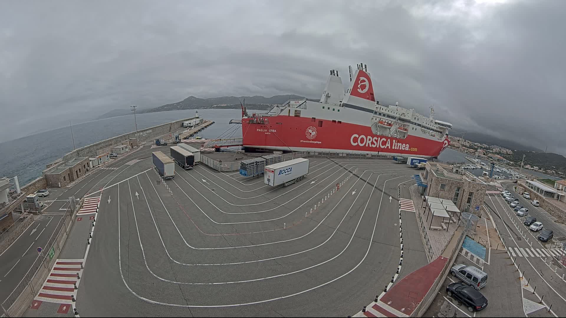 A large red and white ferry is docked at a port on an overcast day, with several trucks and cars in the surrounding area.
