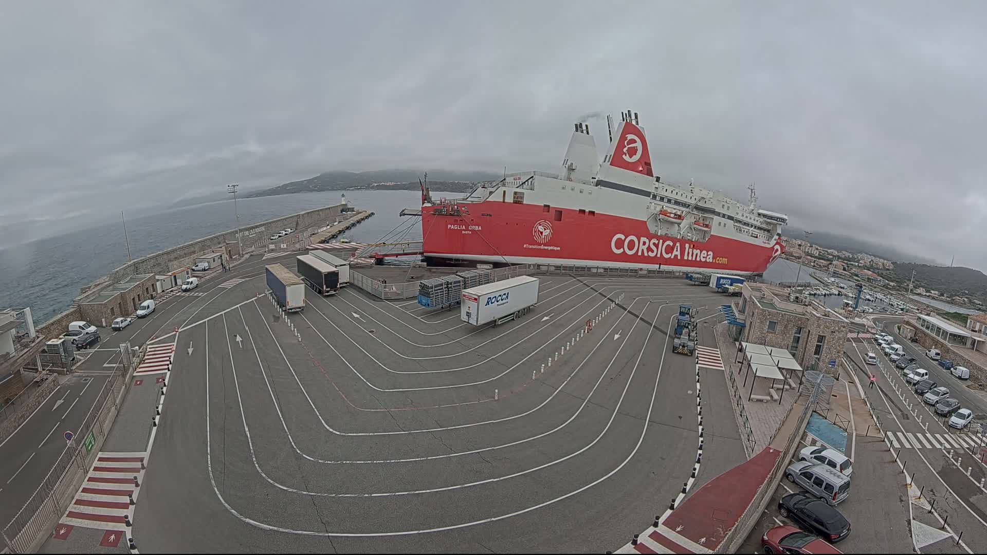 A large red and white ferry is docked at a port on an overcast day, with several vehicles parked nearby and on the ship.