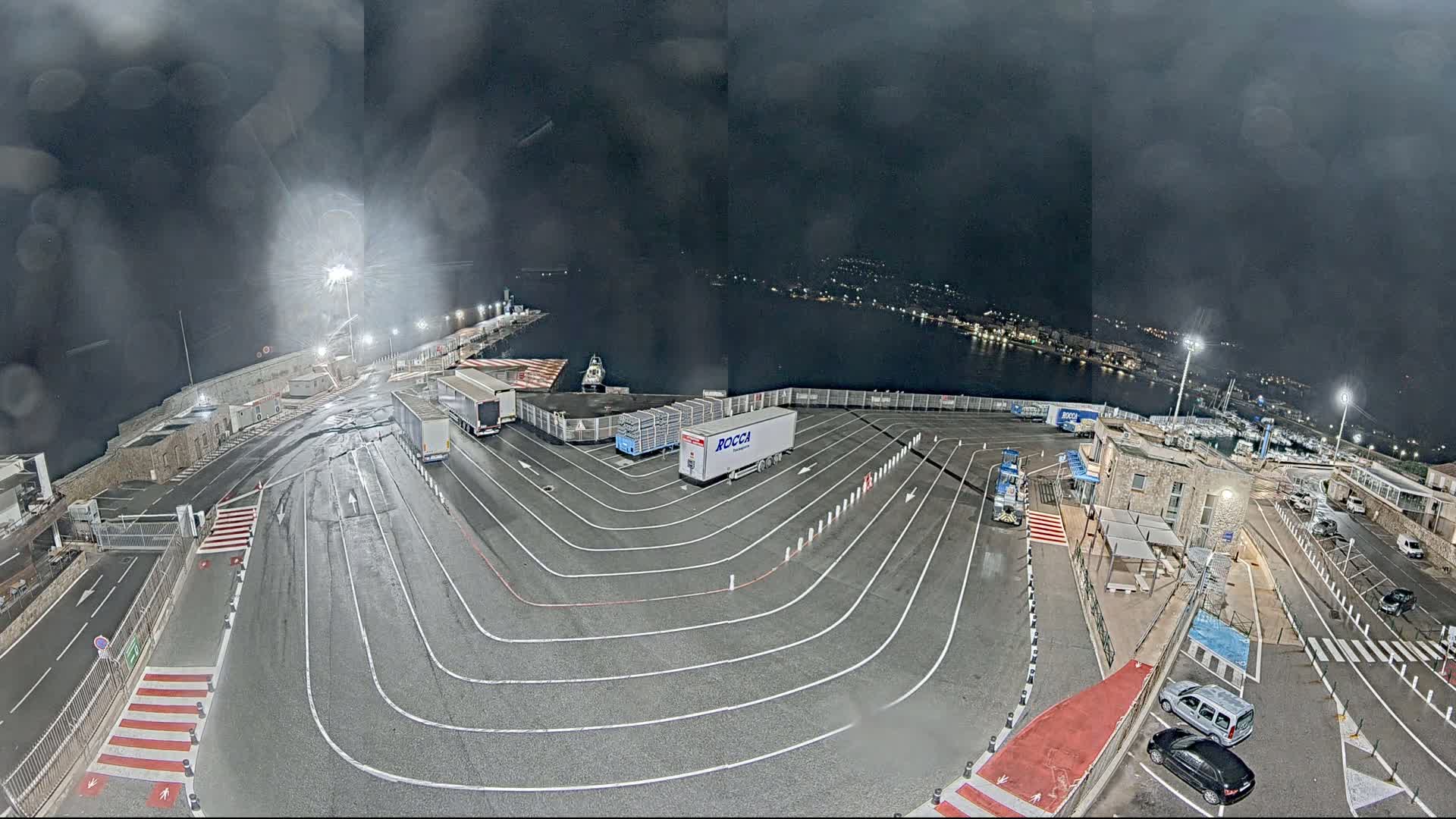 A nighttime view of a port area with several trucks parked in designated lanes under a dark, possibly rainy sky.