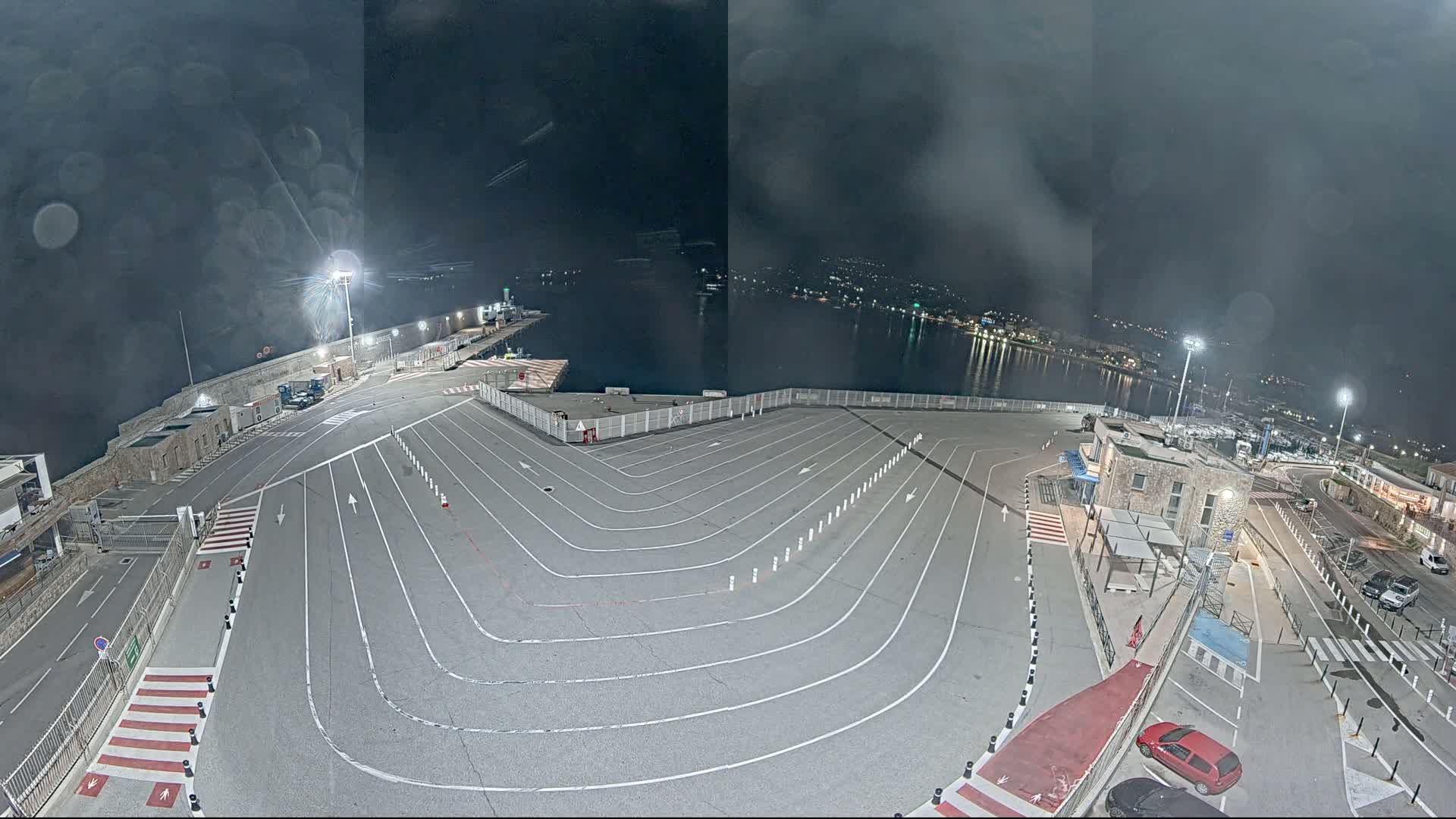 An empty, marked parking lot at a harbor at night under a cloudy sky overlooks a dark body of water with lights visible in the distance.