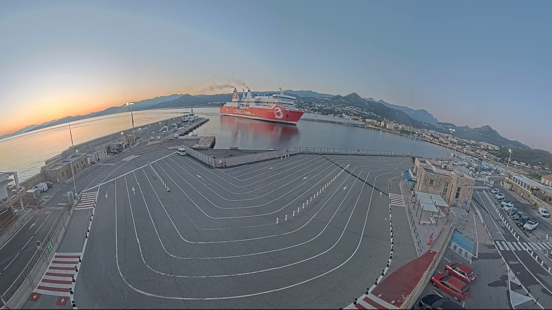 A large red ferry is docked at a harbor at sunrise, with a large paved area for vehicles leading down to the water.
