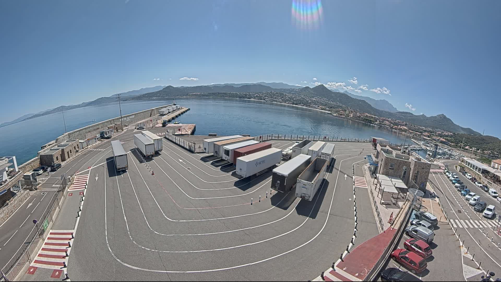 A sunny day reveals a harbor filled with several semi-trucks parked in a designated area, near a body of water with mountains in the background.