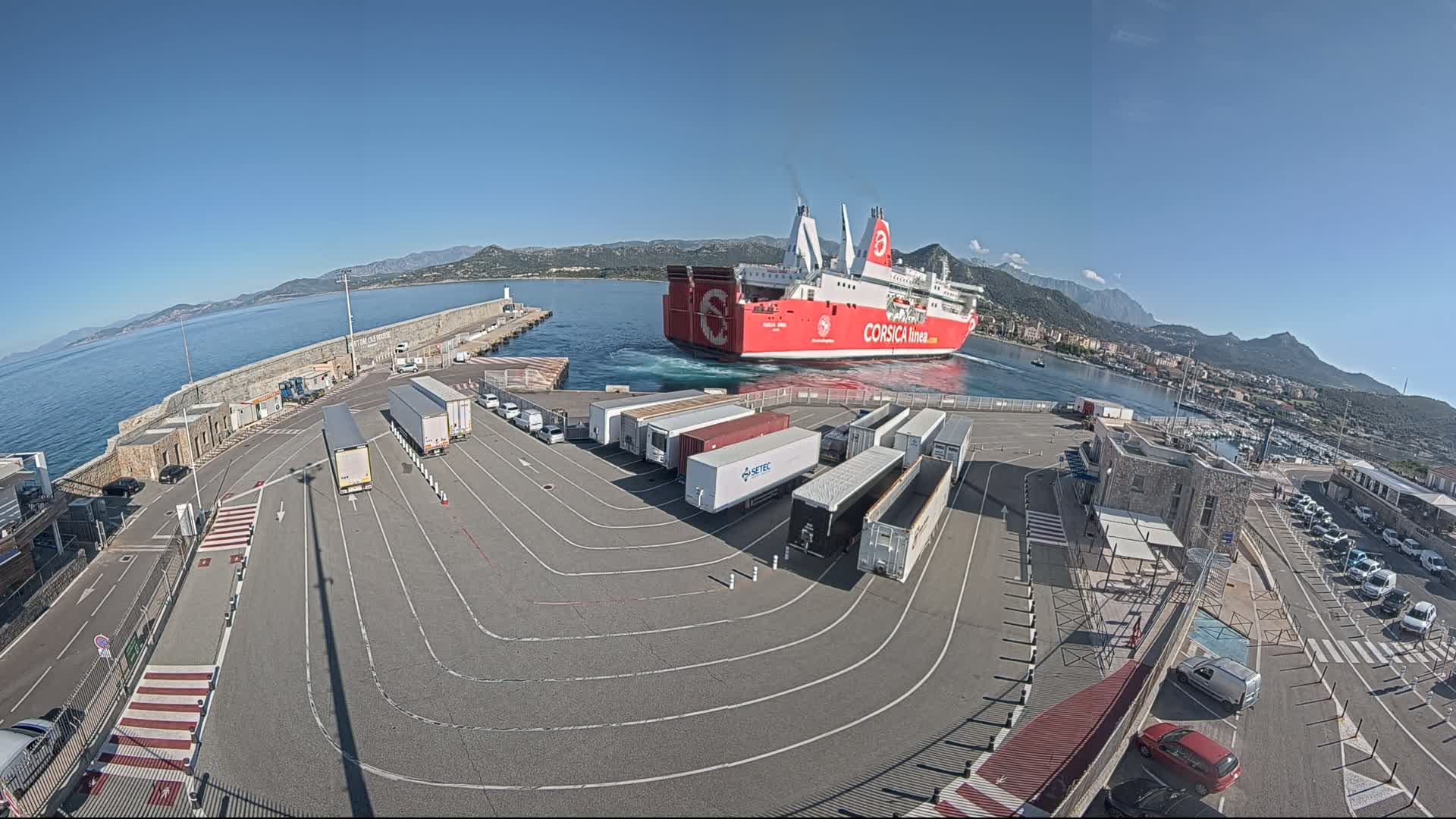 A large red ferry is arriving at a port on a clear, sunny day, with numerous trucks and cars parked in the port's parking lot.