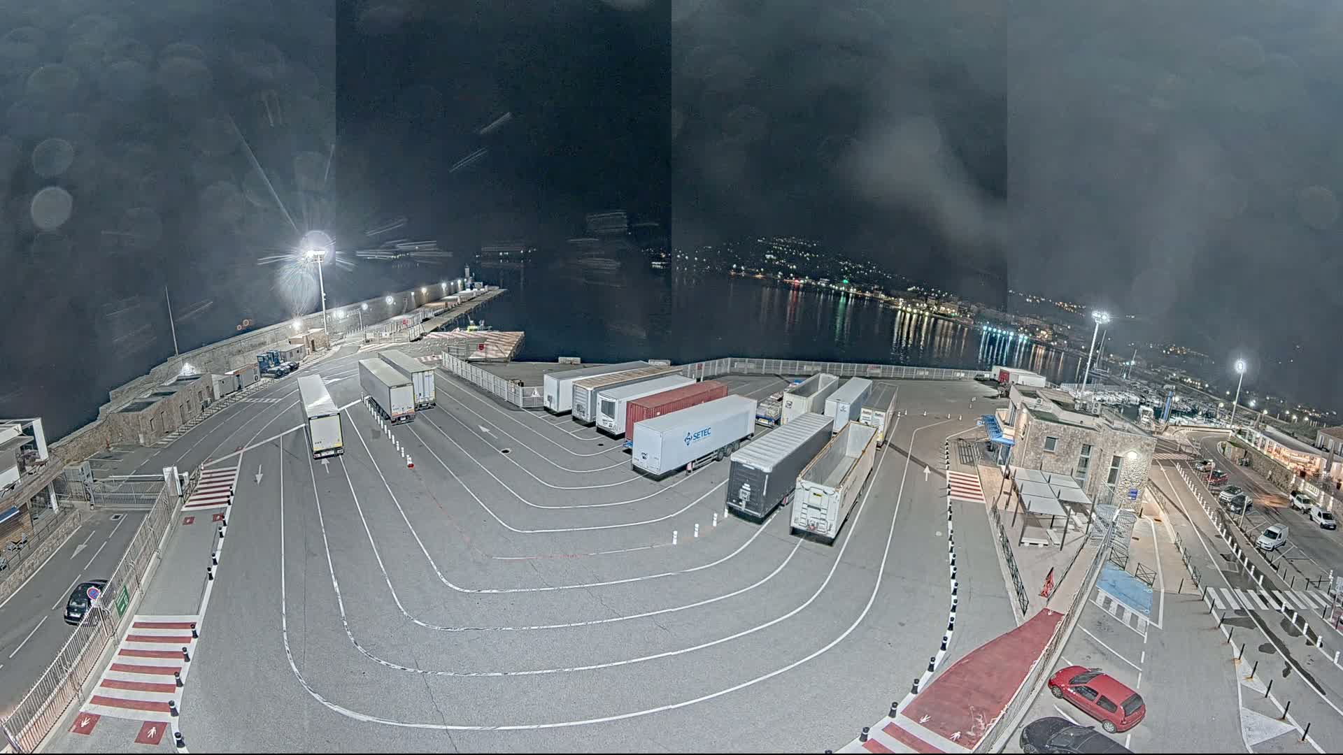 A nighttime aerial view of a port area shows several semi-trailer trucks parked in designated lanes, near a calm body of water with lights visible across the shore under a dark, cloudy sky.