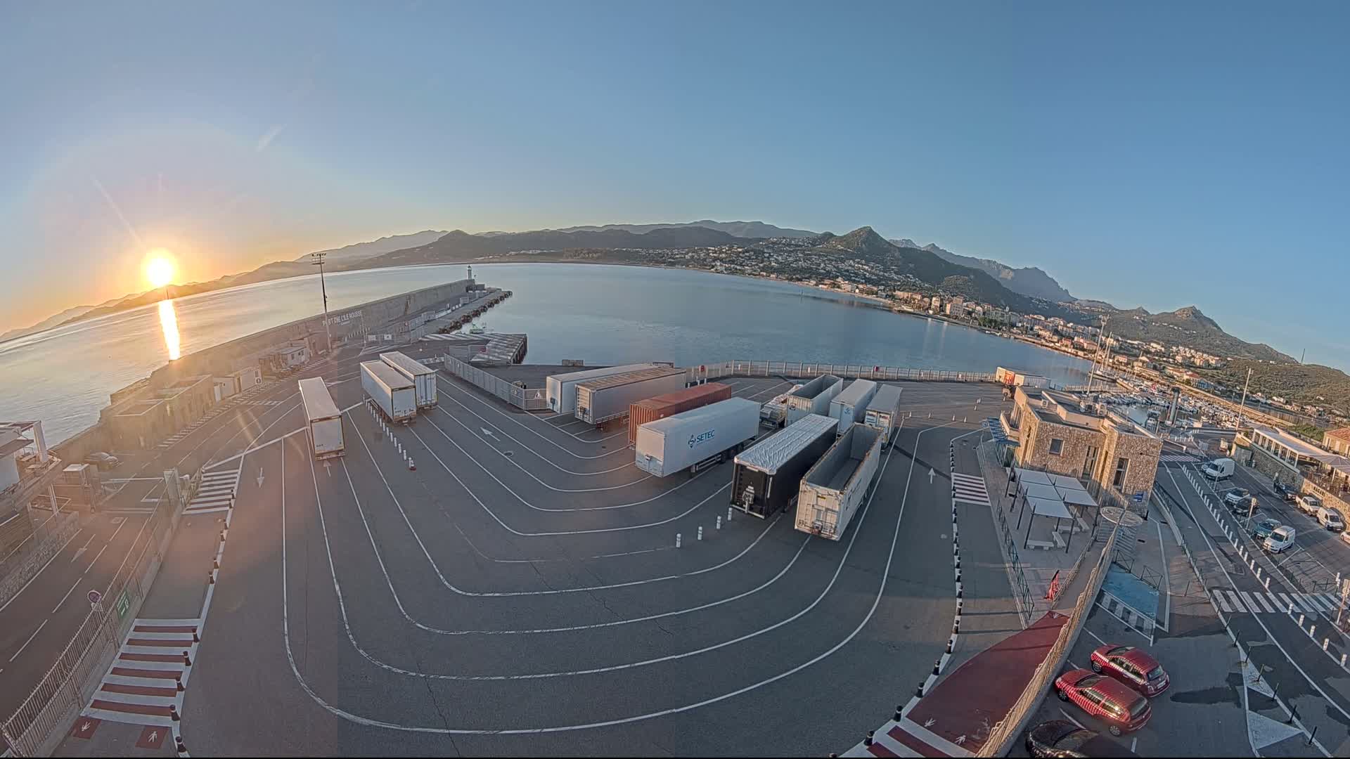 A sunrise illuminates a harbor filled with several semi-trailer trucks parked in a paved lot beside a calm body of water, with mountains visible in the background.