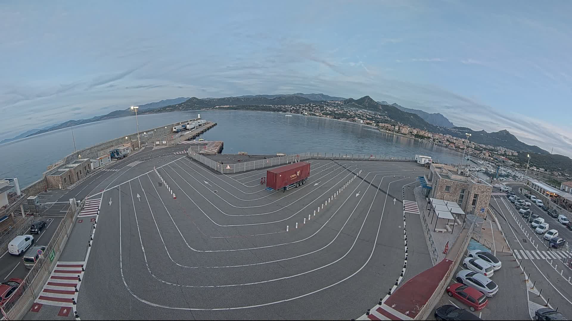 A large, empty paved area with marked lanes and a single red shipping container is situated at a harbor, overlooking calm water and distant mountains under a clear, dusky sky.