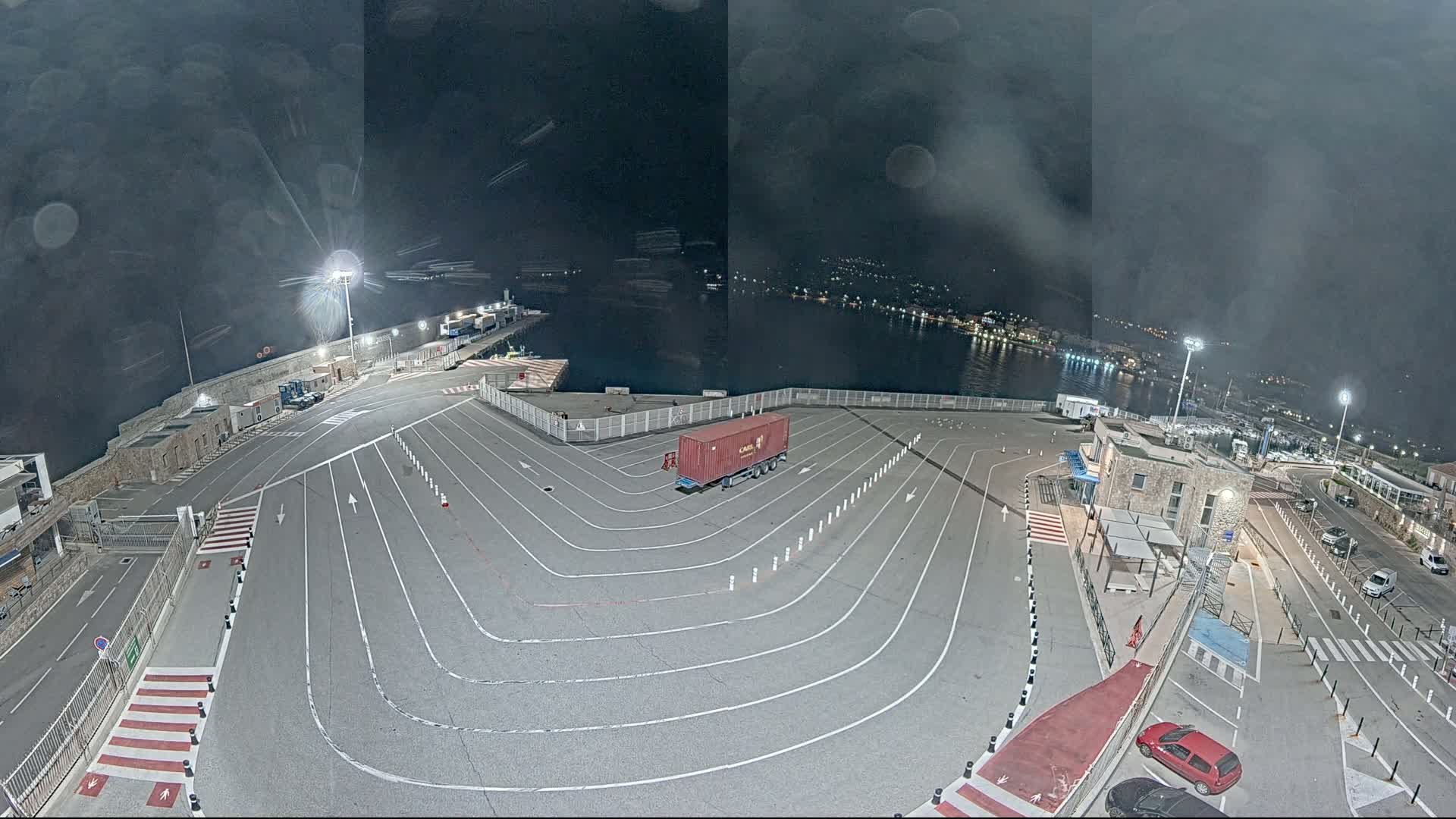 A nighttime aerial view of a port area with a large, empty paved lot marked with driving lanes, a single shipping container, and a few cars parked nearby, under a dark, cloudy sky.