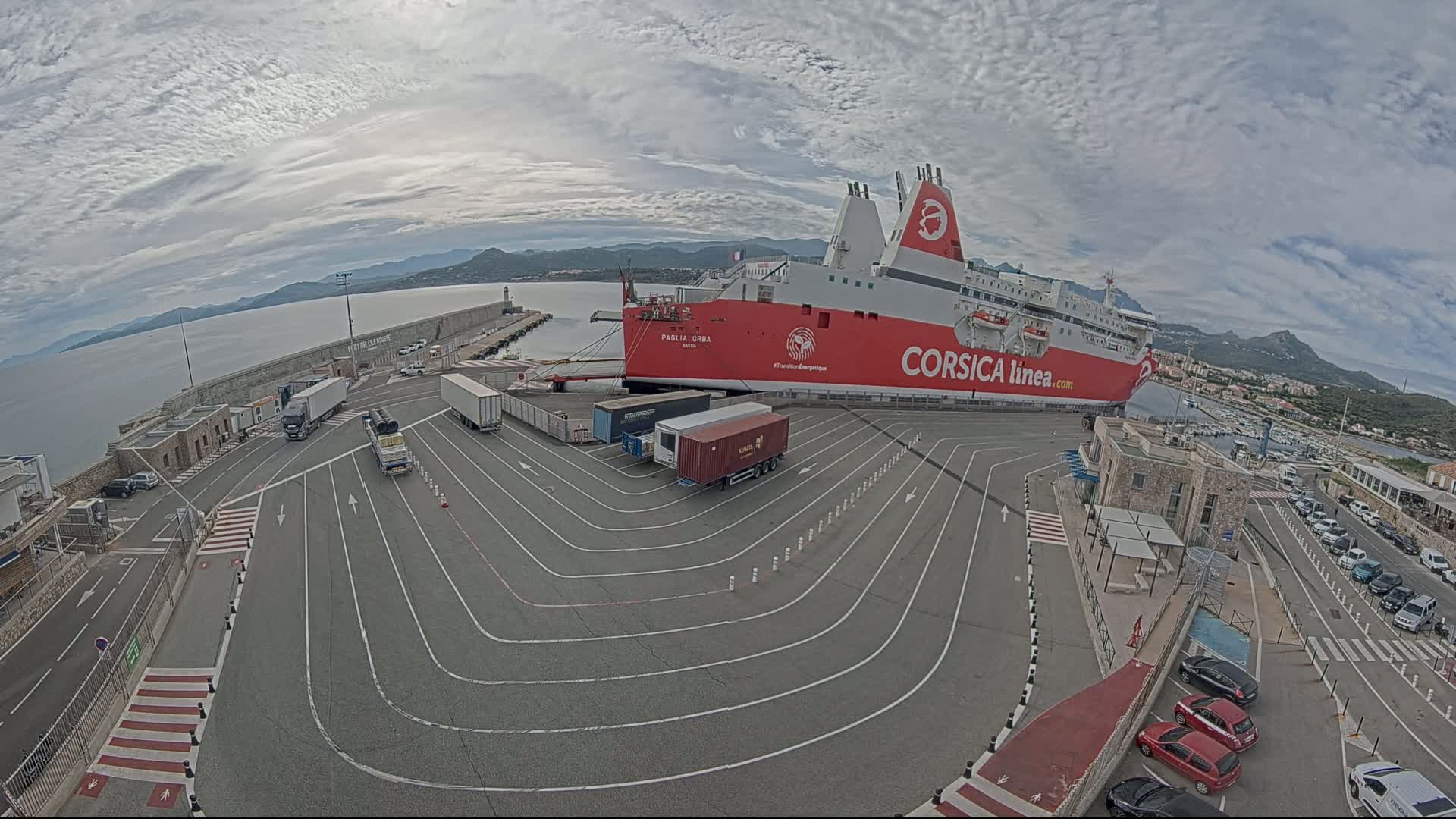 A large red and white ferry is docked at a port on a partly cloudy day, with several trucks and cars parked nearby in a designated area.