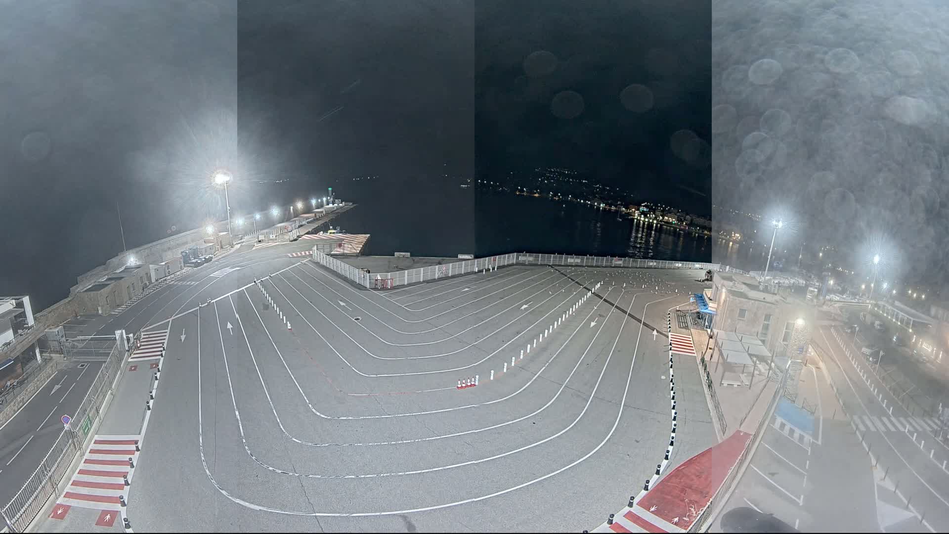 An empty, nighttime ferry terminal with marked lanes is situated beside a body of water under a hazy, dark sky.