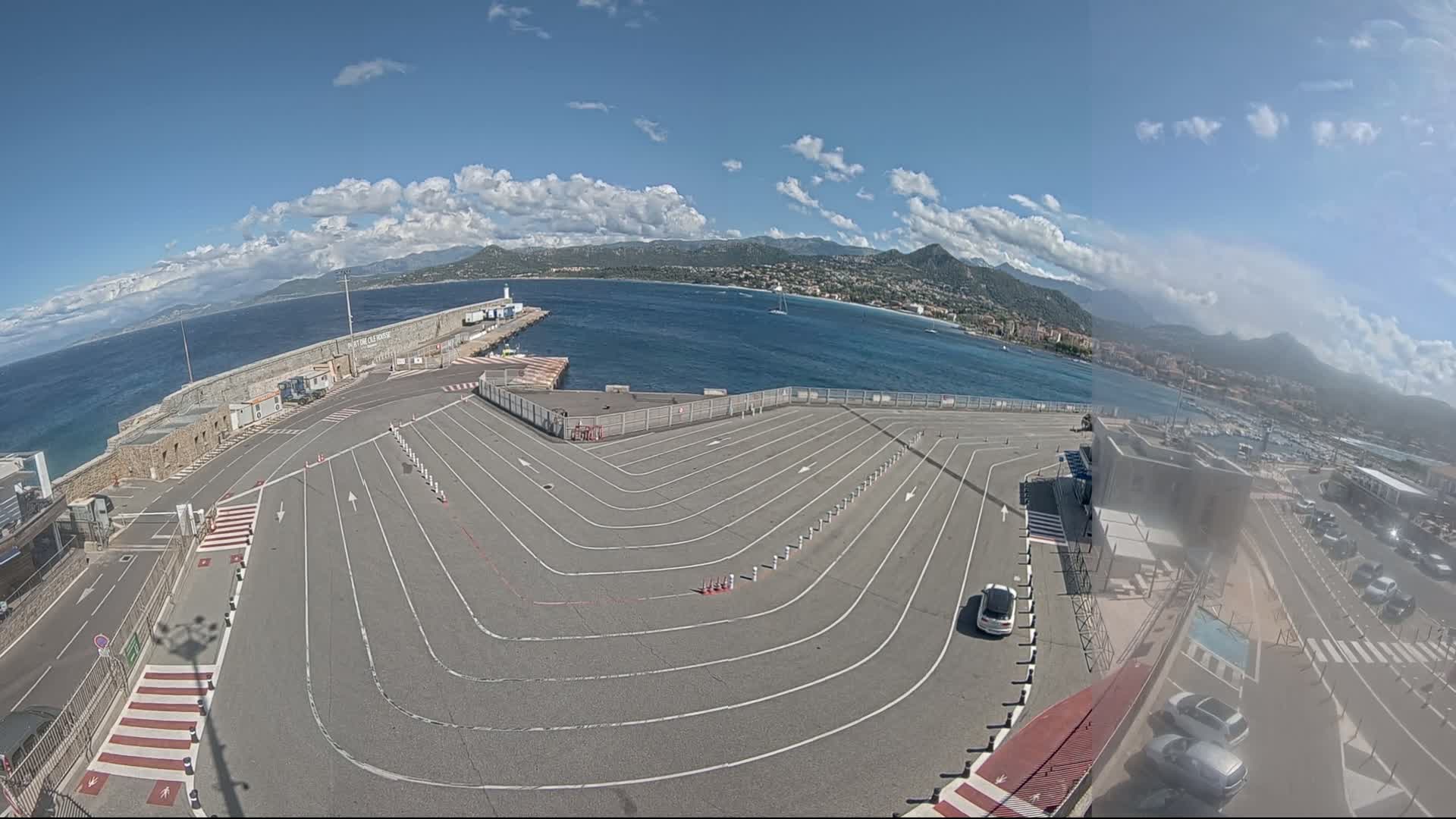 A mostly empty, curved vehicle staging area at a port overlooks a calm sea under a partly cloudy sky, with mountains visible in the distance.