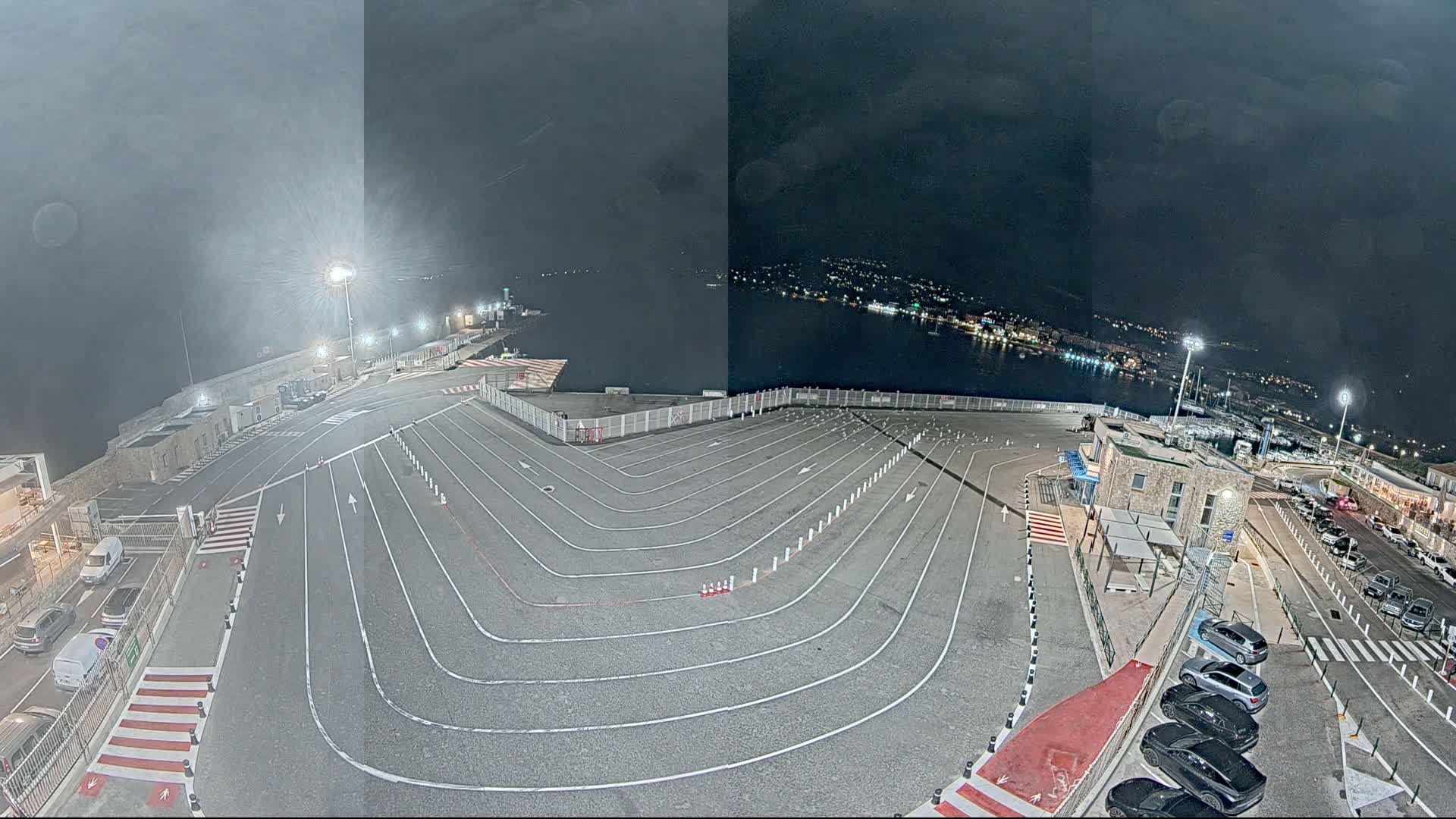 An empty, lined parking lot at night overlooks a dark body of water with a town lit up in the distance under a cloudy sky.