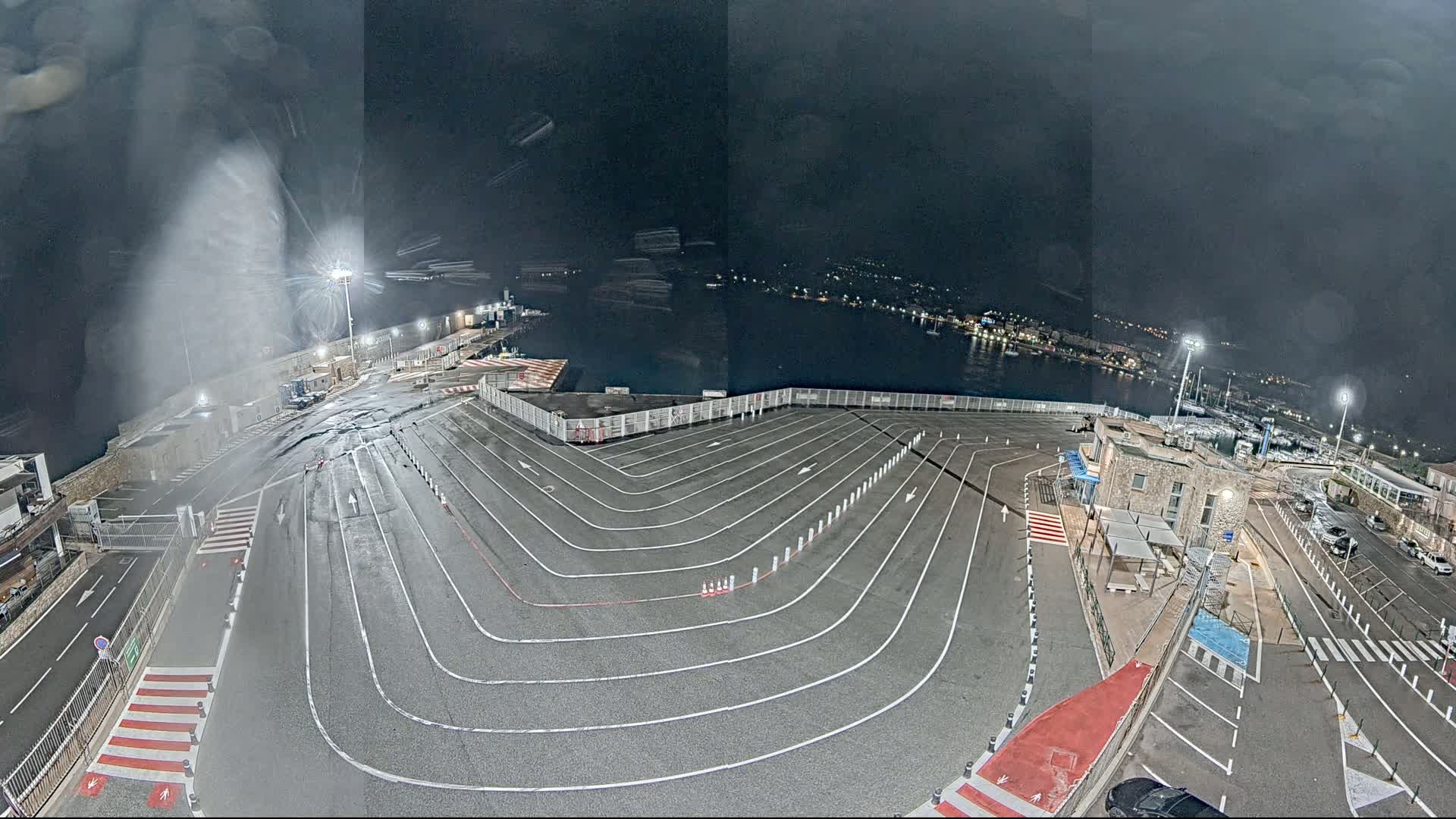 A nighttime aerial view shows a wet, empty ferry terminal with marked lanes curving toward a body of water under a dark, cloudy sky.