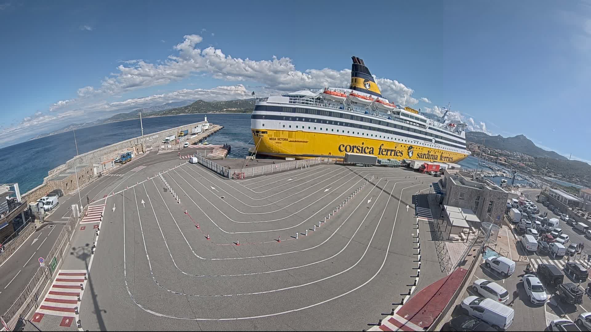 A large yellow and white ferry is docked at a port under a partly cloudy blue sky, with numerous cars parked nearby.