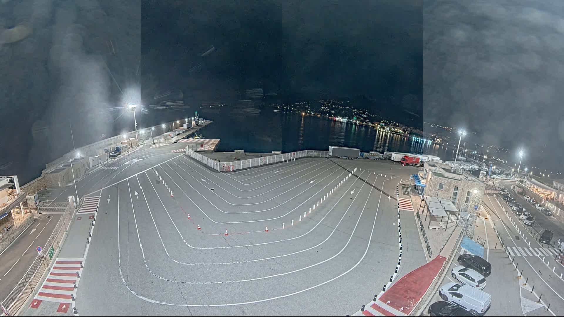 A nighttime aerial view of a harbor's empty vehicle staging area with a calm body of water and a town visible in the distance under a cloudy sky.