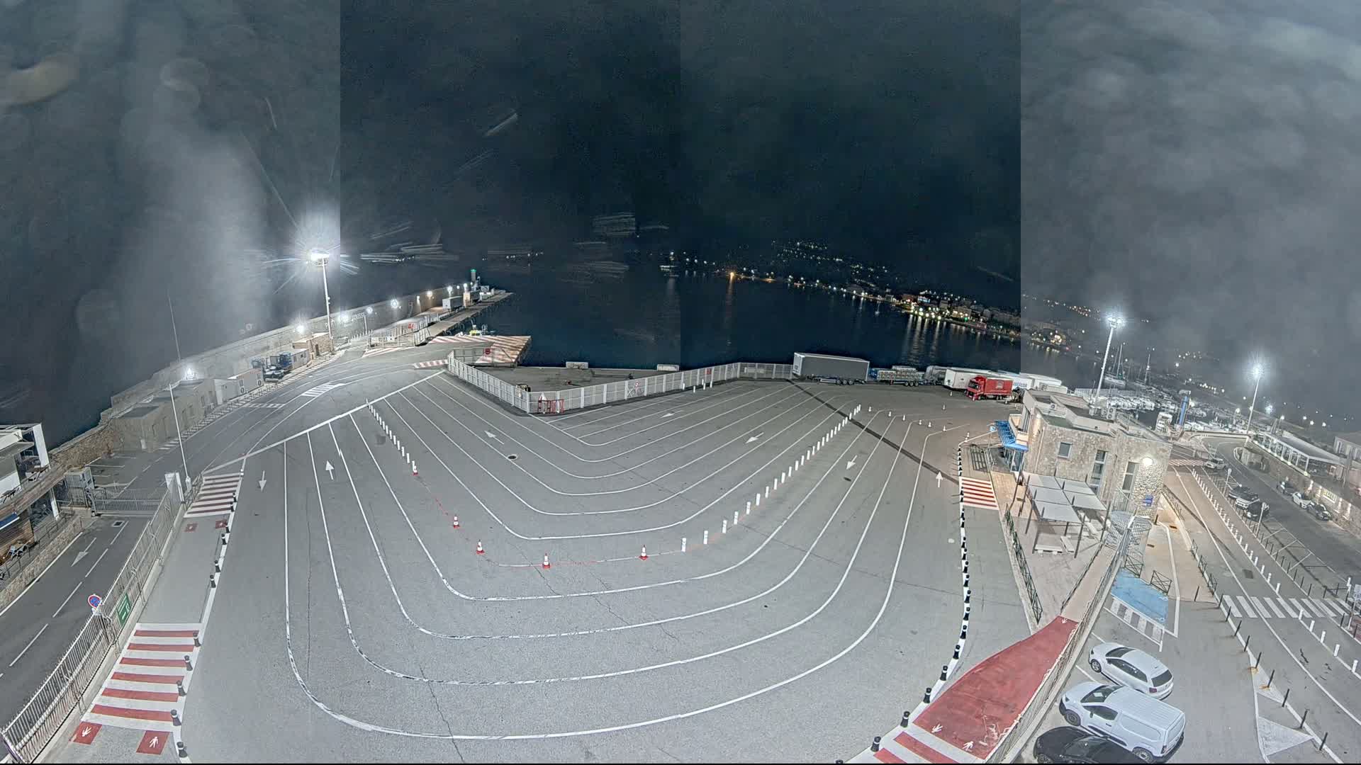 A nighttime, high-angle view of a harbor's vehicle staging area with a body of water and distant lights in the background.