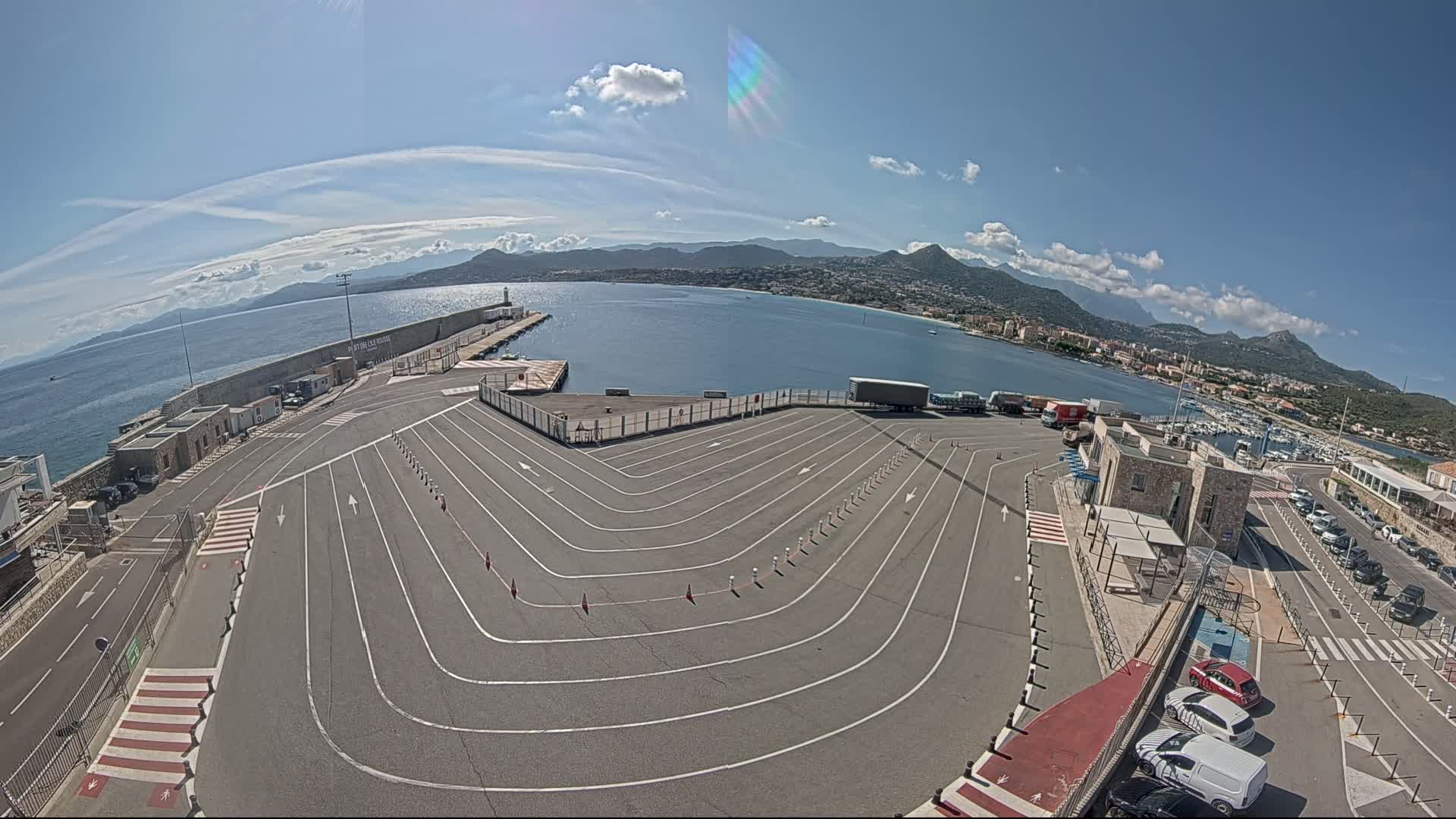 A port with a large, empty vehicle queuing area overlooks a calm bay under a mostly sunny sky, mountains visible in the distance.