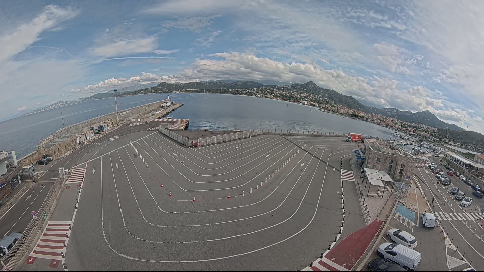 A mostly sunny day shows a large paved area with marked driving lanes leading to a harbor, a body of water, and distant mountains.