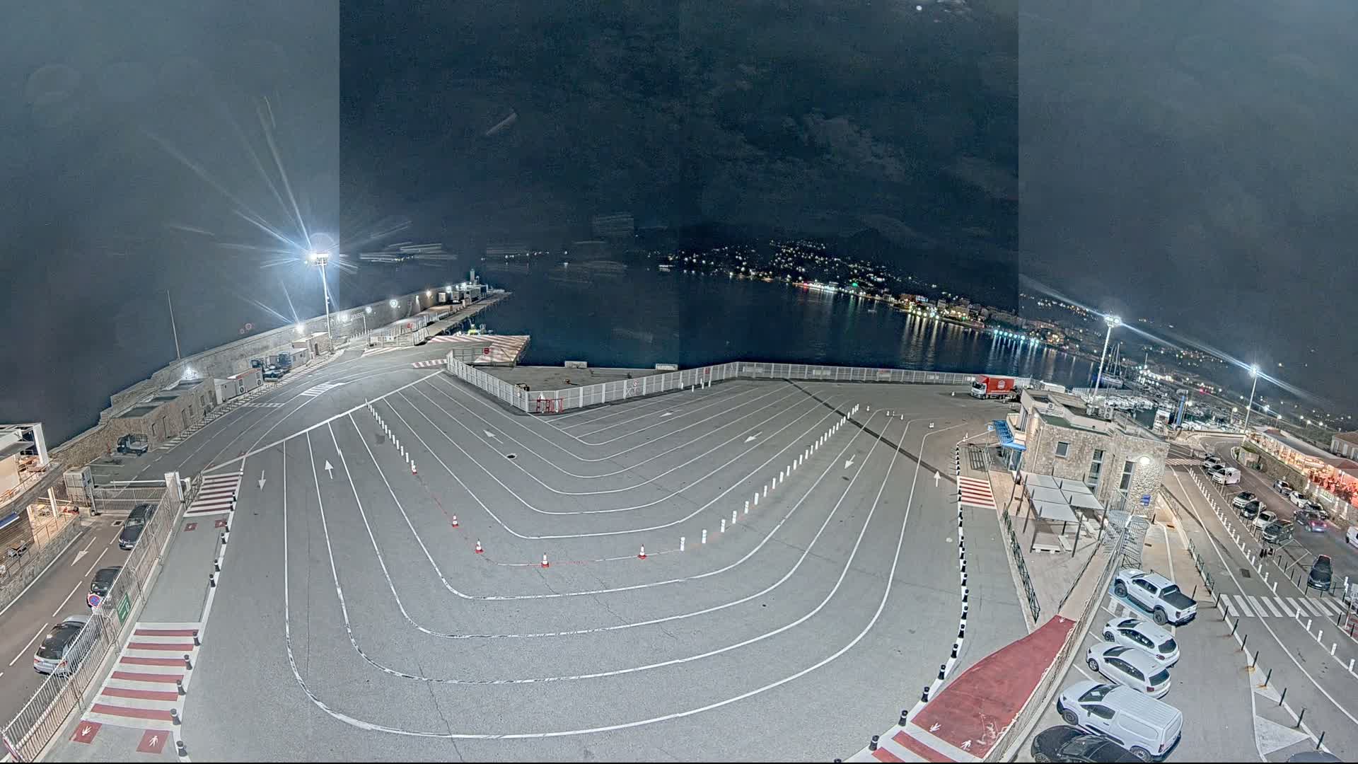 A nighttime aerial view shows a harbor's empty vehicle staging area with marked lanes, a few parked cars, and a calm body of water with lights visible on the opposite shore.