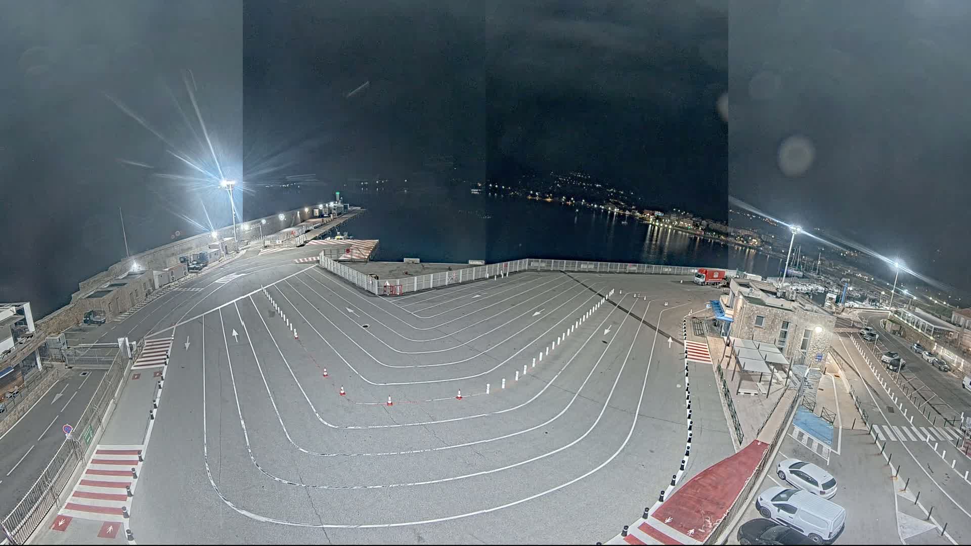An empty, marked parking lot at night next to a calm harbor, illuminated by bright lights.