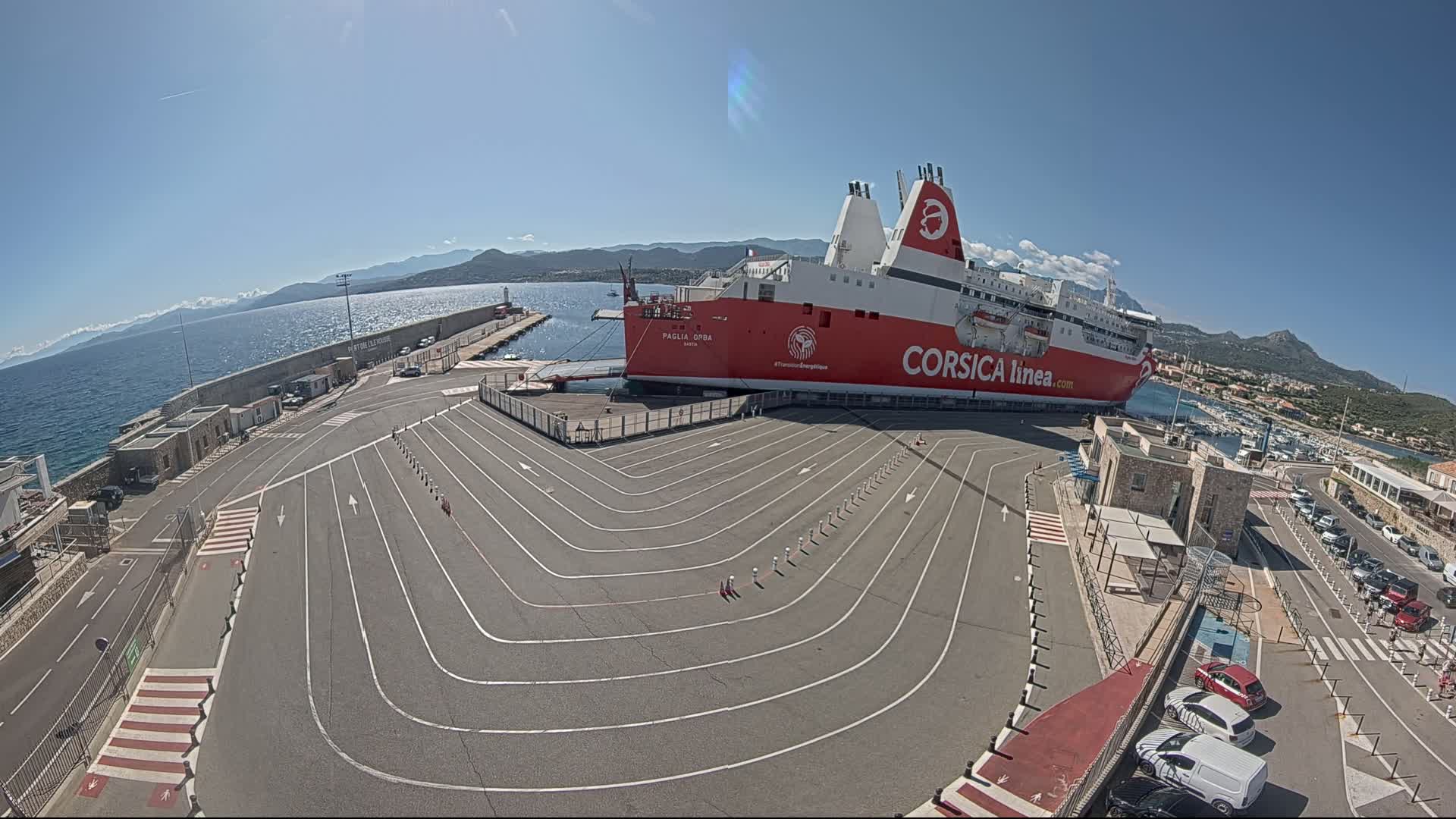 A large red ferry is docked at a port on a sunny day, with a paved area marked with lines for vehicle queuing.