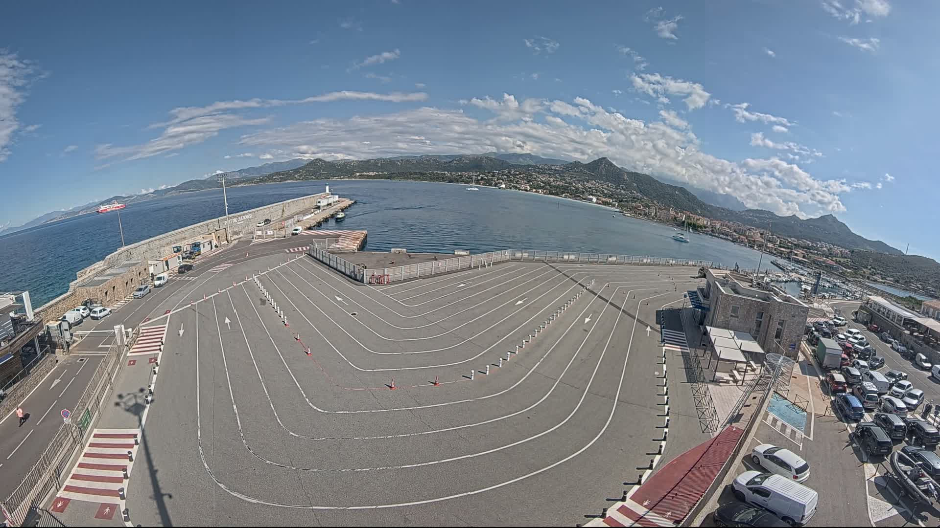 A large, empty ferry terminal with marked lanes curves towards a body of water under a mostly sunny sky with some clouds, mountains in the distance, and cars parked in a lot nearby.