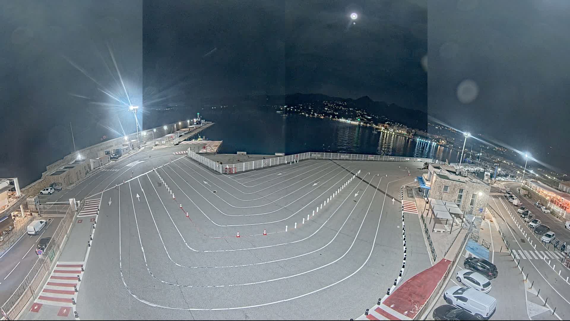 An empty, marked, nighttime parking lot overlooks a calm harbor with a moonlit city in the distance.