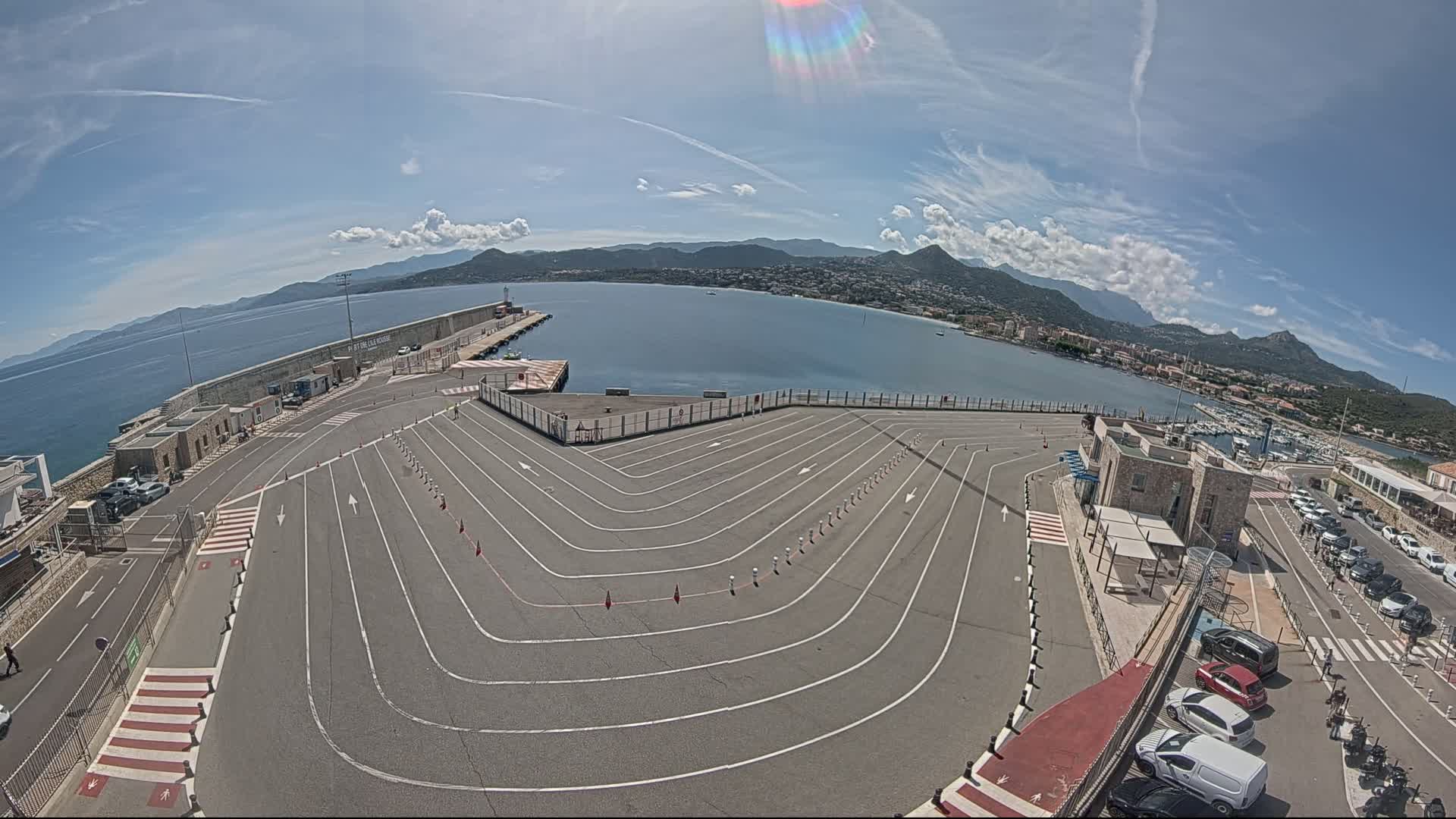 A mostly empty, curved vehicle queuing area at a harbor overlooks a calm sea and mountainous landscape under a partly sunny sky.