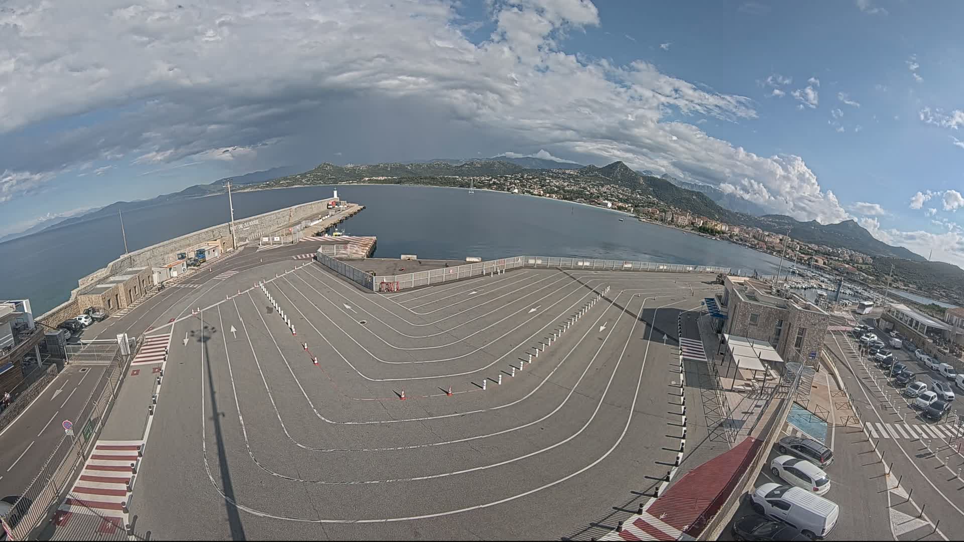 An empty, marked parking lot overlooks a calm bay with mountains in the distance under a partly cloudy sky.