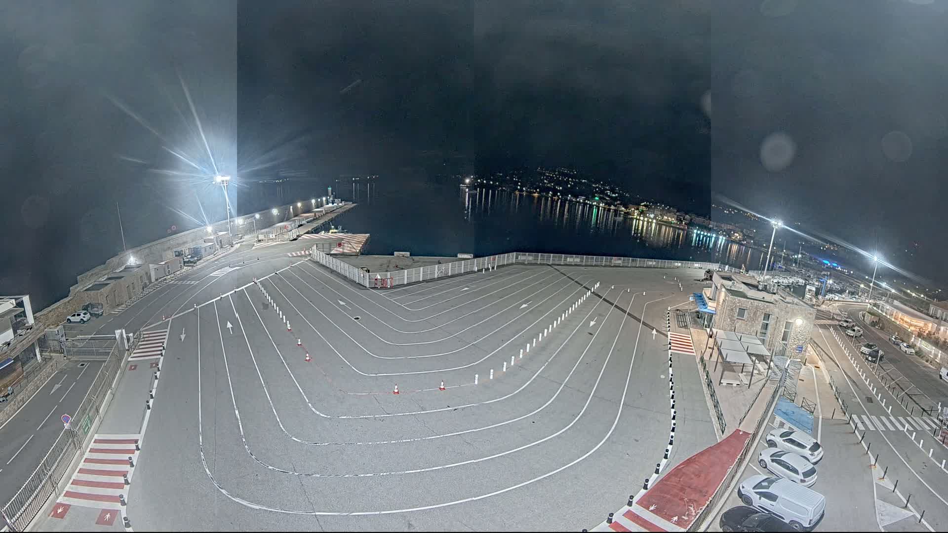 An empty, marked parking lot at night overlooks a calm, dark body of water reflecting city lights under a cloudy sky.