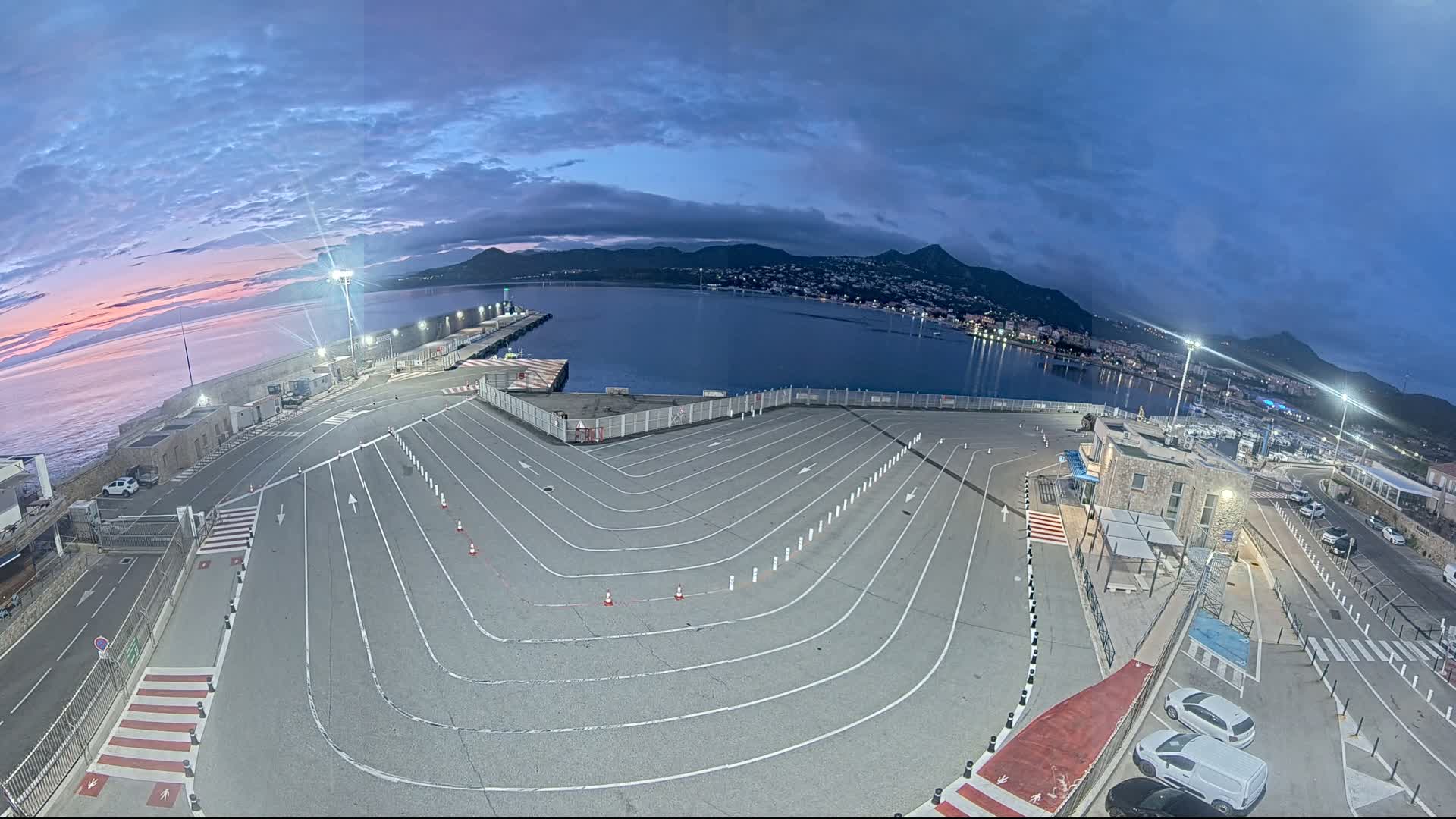 An empty, striped parking lot at a harbor overlooks calm water and a town nestled against mountains under a twilight sky.