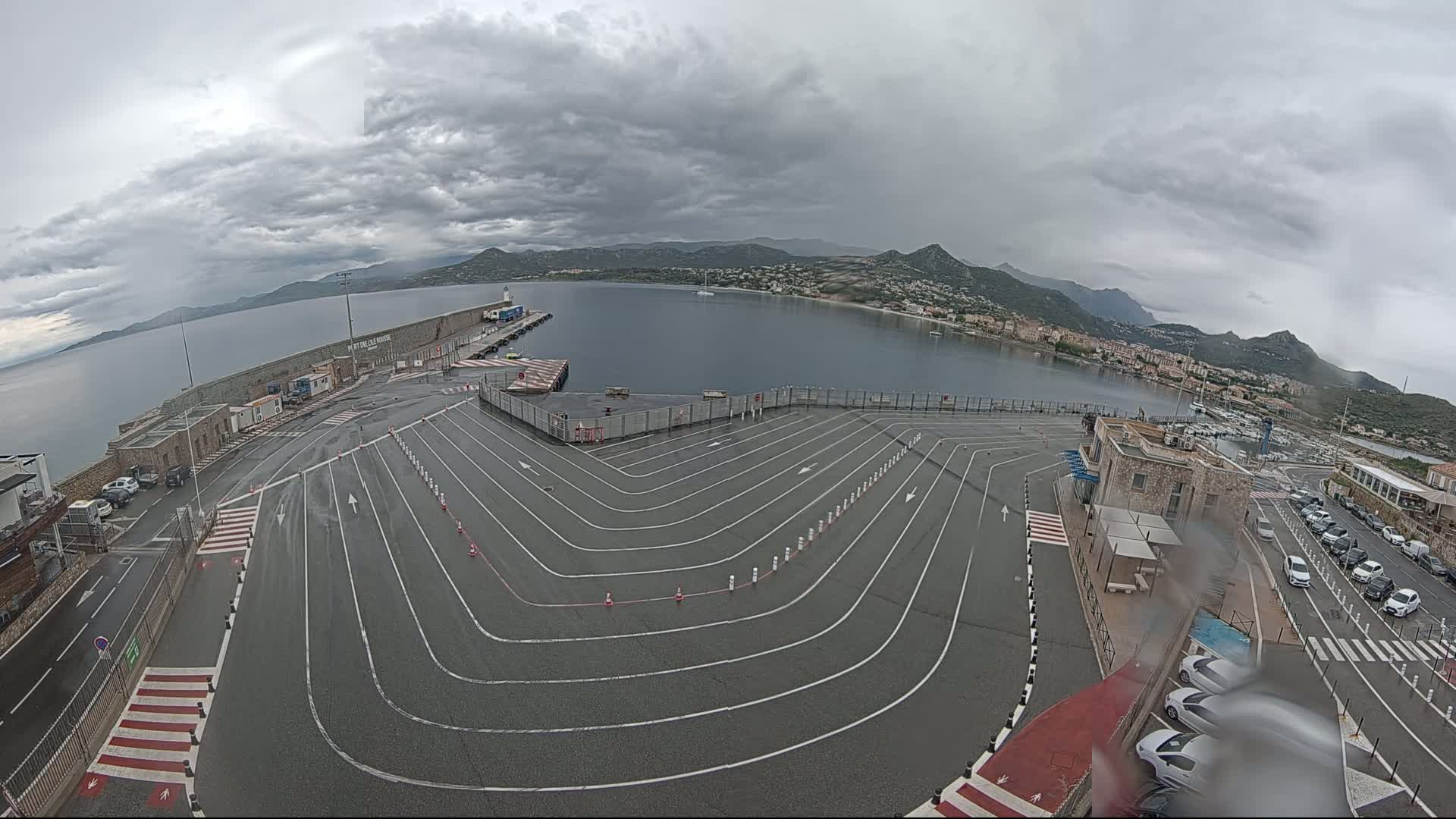 A wet, paved ferry terminal area with marked lanes curves toward a calm bay under a cloudy sky, mountains visible in the background.