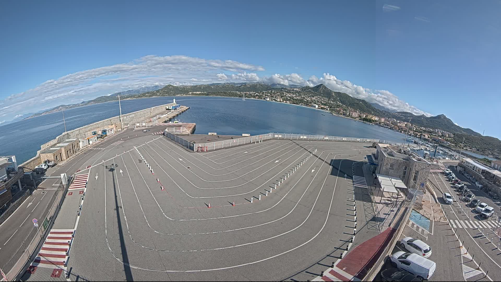 A large, empty vehicle staging area curves around a harbor on a sunny day with partly cloudy skies and mountains in the background.