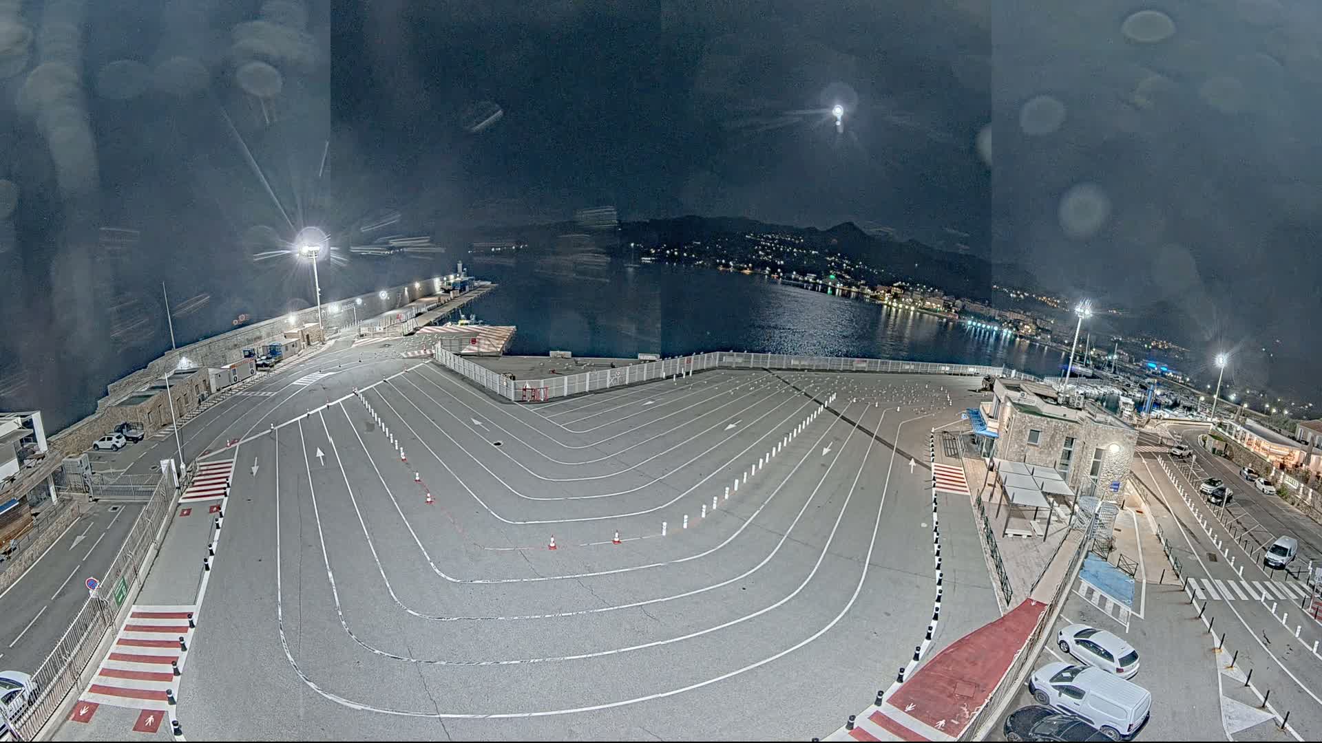 A nighttime aerial view of an empty, marked parking lot at a harbor, with a moon in the sky and lights illuminating the scene.